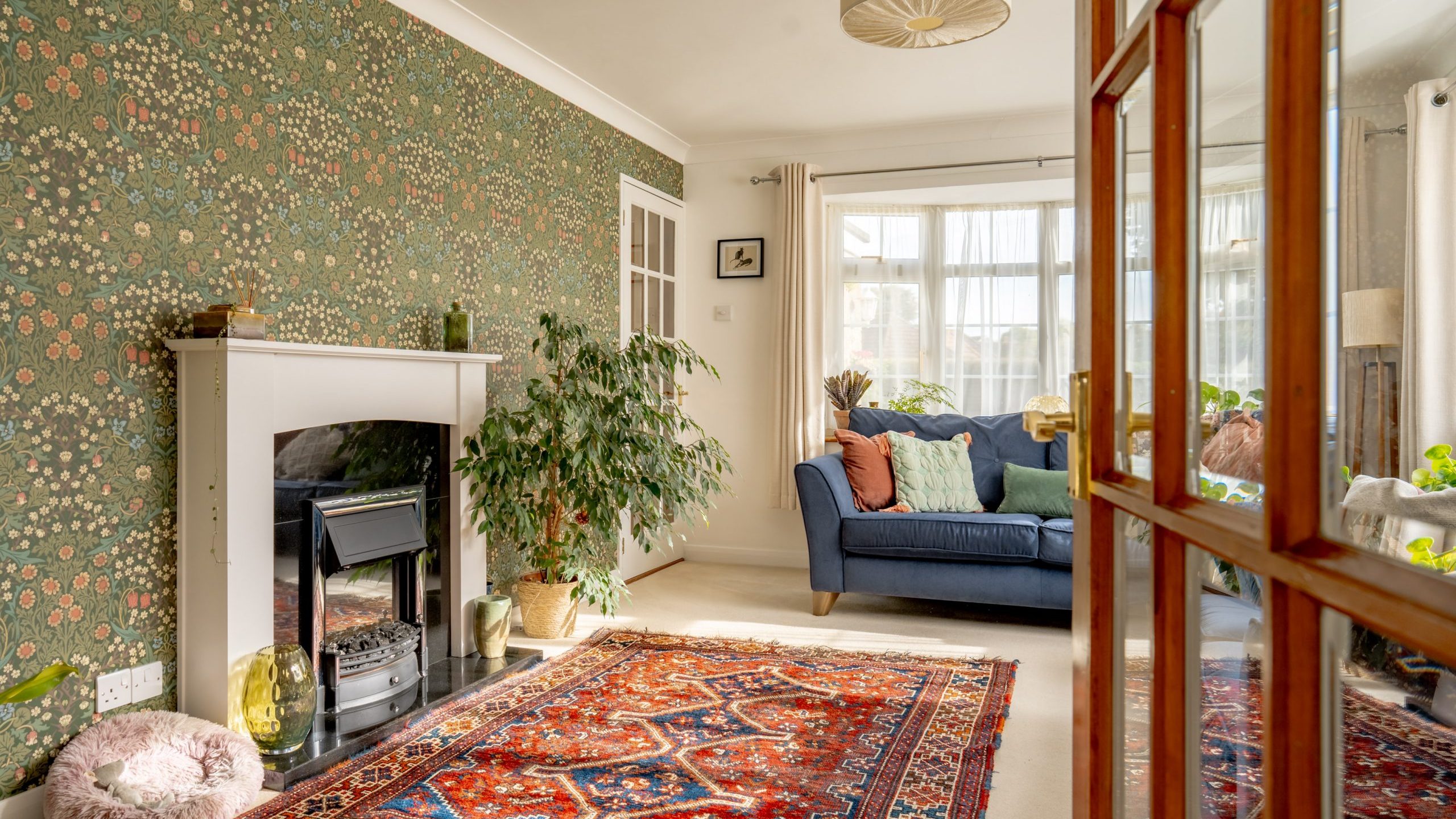 A cozy living room with a colorful Persian rug, a blue sofa adorned with cushions, and a green potted plant next to a white fireplace. The feature wall is covered in floral wallpaper, and natural light streams in through a large window.