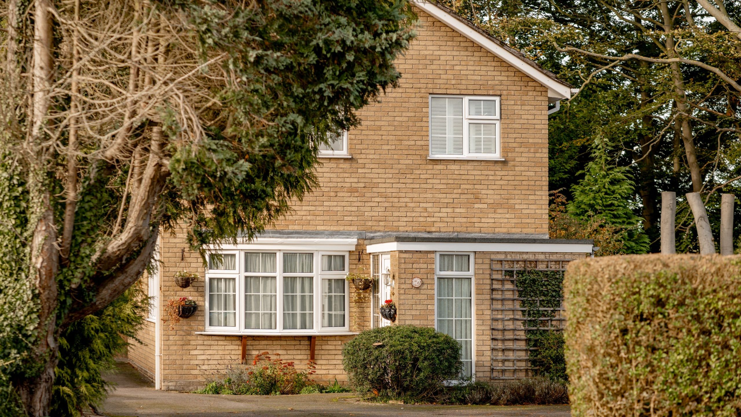 A two-story brick house with a chimney, surrounded by trees and shrubs. It has a bay window on the ground floor and a driveway leading to the entrance. The garden is well-maintained, creating a cozy, suburban feel.