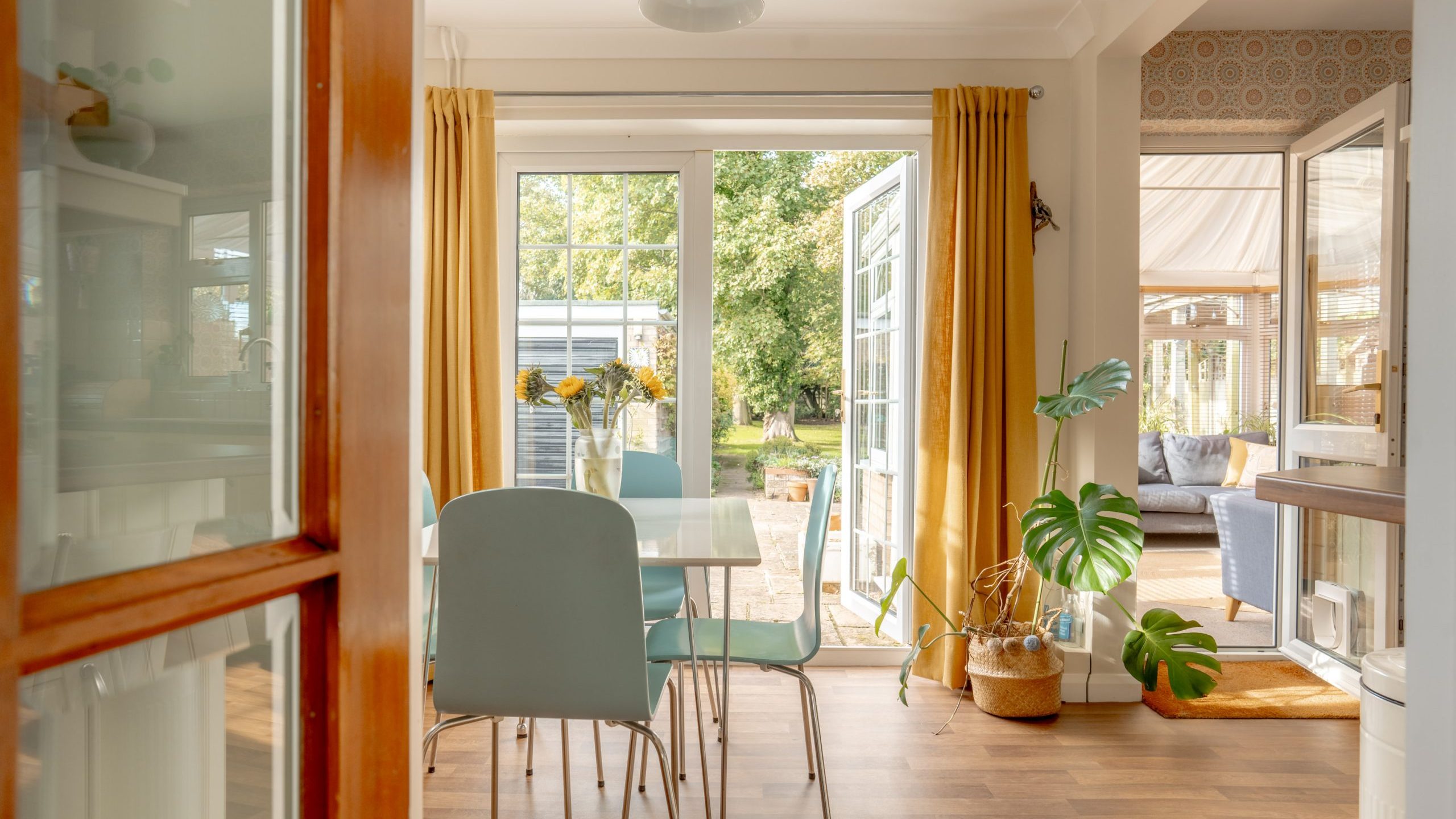 Bright dining room with light blue chairs and a white table near open glass doors leading to a garden. Yellow curtains frame the doors. A plant sits in the corner, and light streams in, creating a warm, inviting atmosphere.