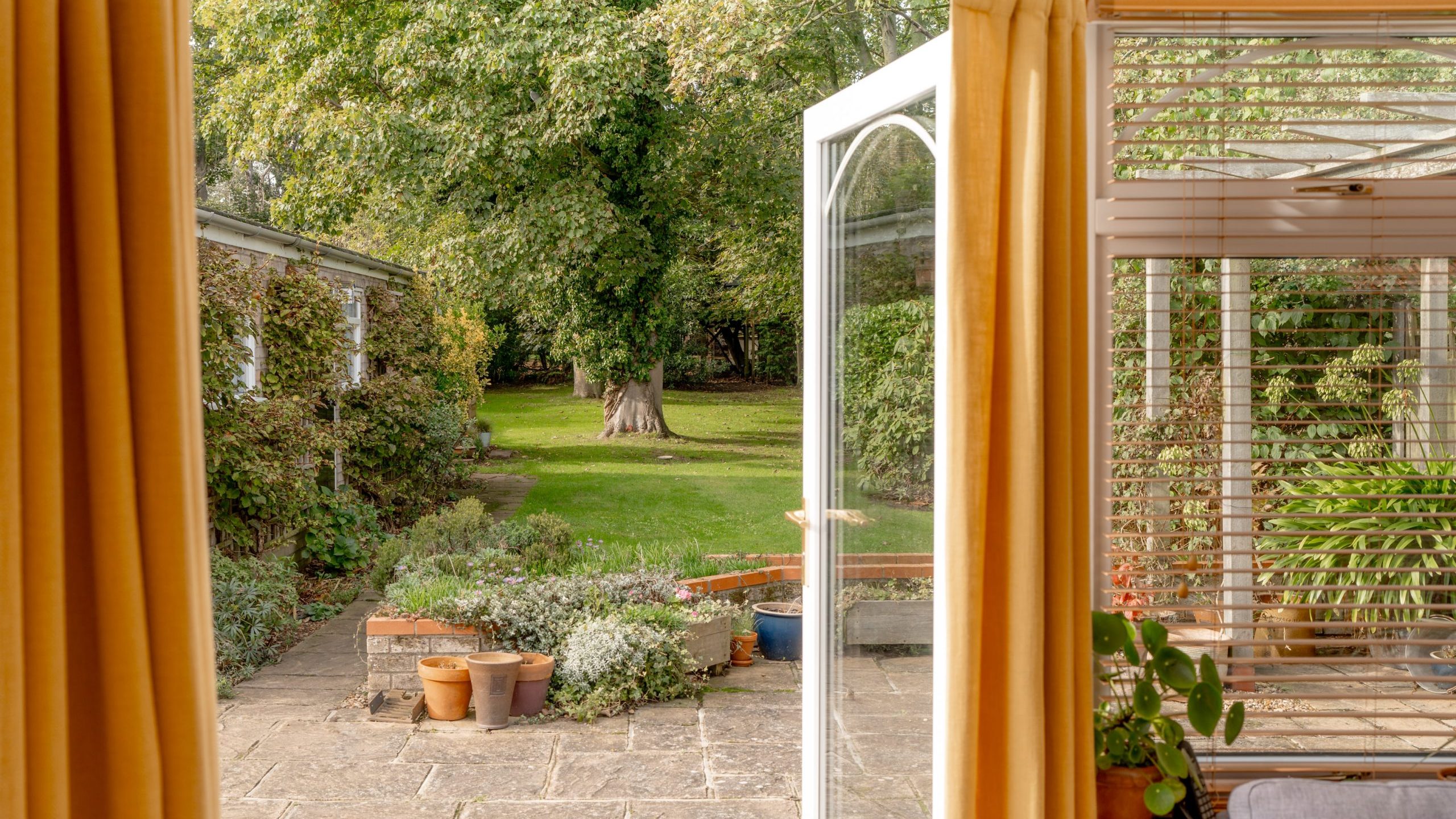 A sunlit garden view from inside a home, featuring an open glass door with yellow curtains on either side. Potted plants sit on a stone patio, leading to a lush green lawn and trees in the background.
