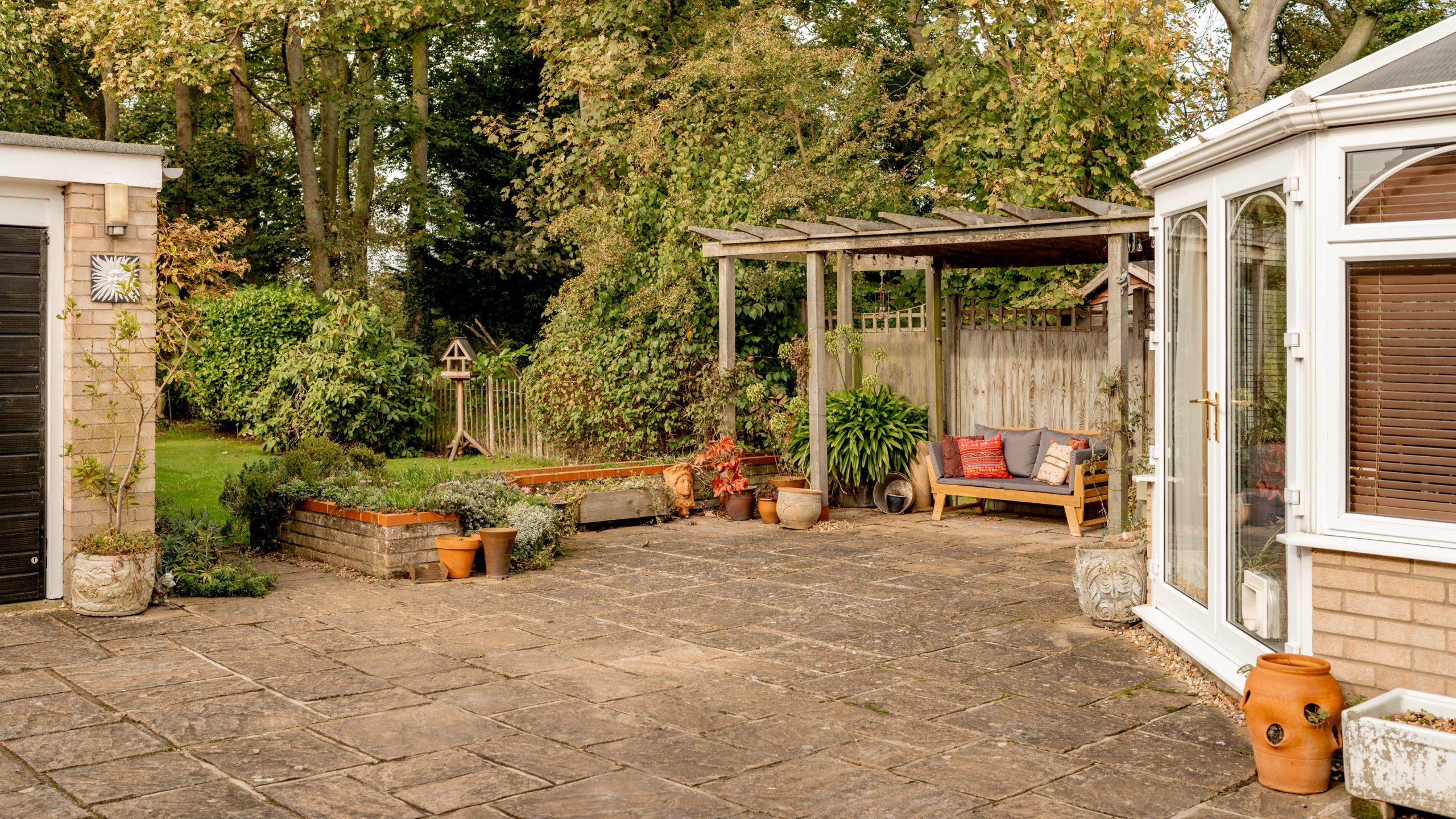 A spacious backyard patio featuring potted plants, a wooden pergola with seating and colorful cushions, and a garden area with trees in the background. A white conservatory and a wall clock are visible on the left.