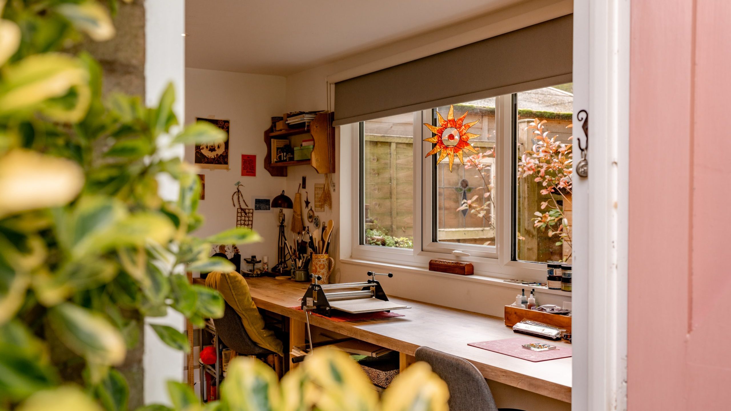 A cozy home office with a long wooden desk by a window, decorated with art supplies and a colorful sun ornament. Two chairs are placed in front of the desk. Greenery frames the doorway, providing a view of a garden outside.