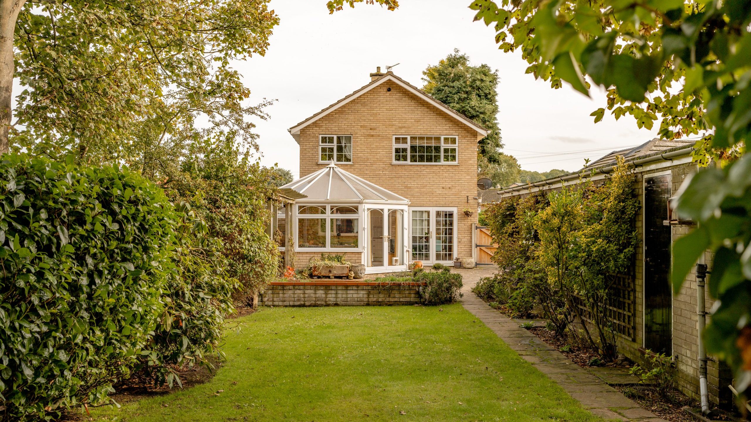 A two-story brick house with a glass conservatory is surrounded by greenery. A neatly maintained lawn leads to the house, bordered by bushes and trees. A paved pathway runs along the right side, and the sky is overcast.