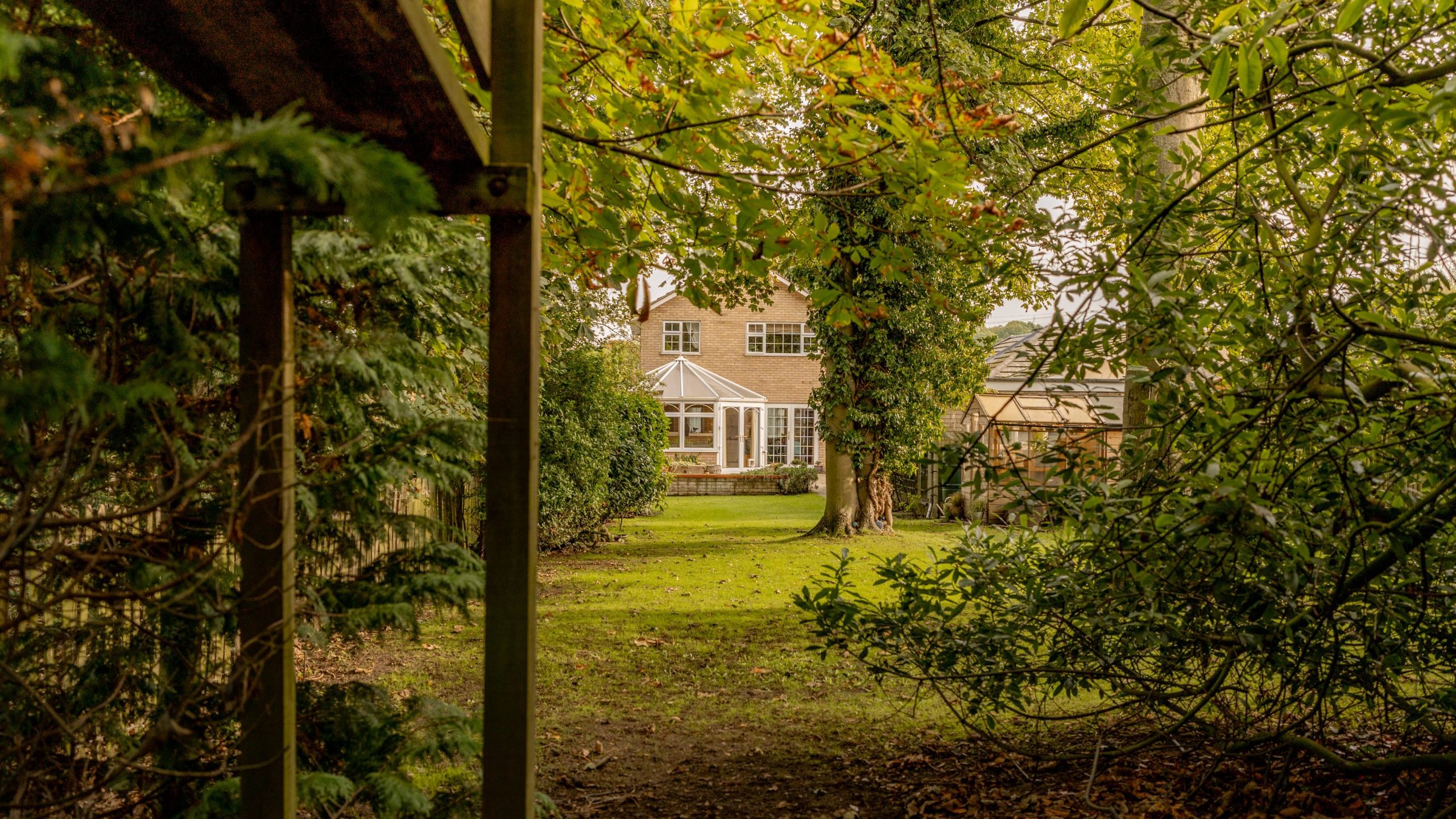 A view of a house with a glass-roofed conservatory, surrounded by lush greenery and trees. The perspective is from a garden pathway bordered by bushes and trees, creating a secluded, tranquil atmosphere.