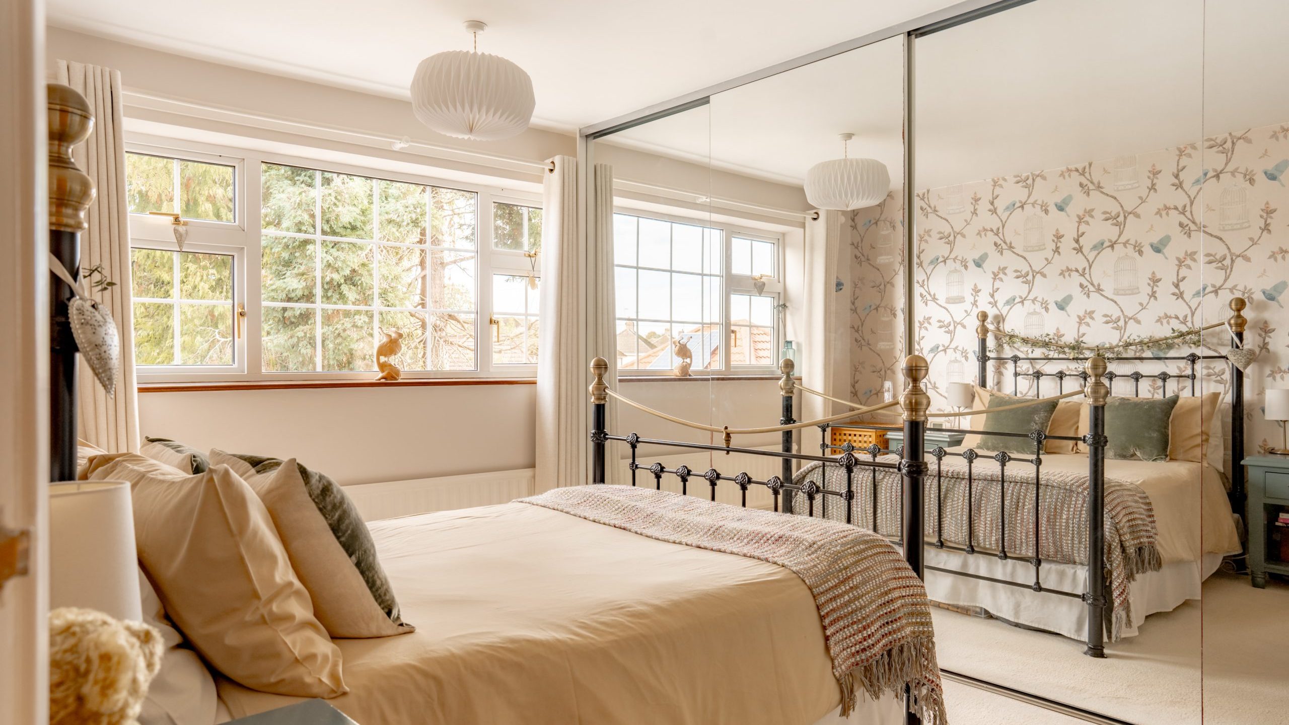 A cozy bedroom with a metal bed, beige bedding, and a plaid throw. Large windows bring in natural light. A mirrored wardrobe reflects floral wallpaper. A teddy bear sits on a side table, and pendant lamps hang from the ceiling.