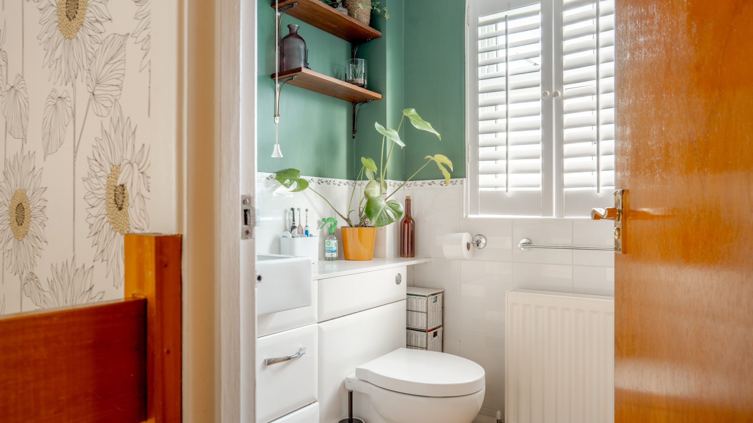 A small bathroom with green walls and white accents features a white toilet and sink with storage cabinets. Plants and toiletries sit on wooden shelves and countertops. A window with shutters lets in light, and a wooden door is partially open.