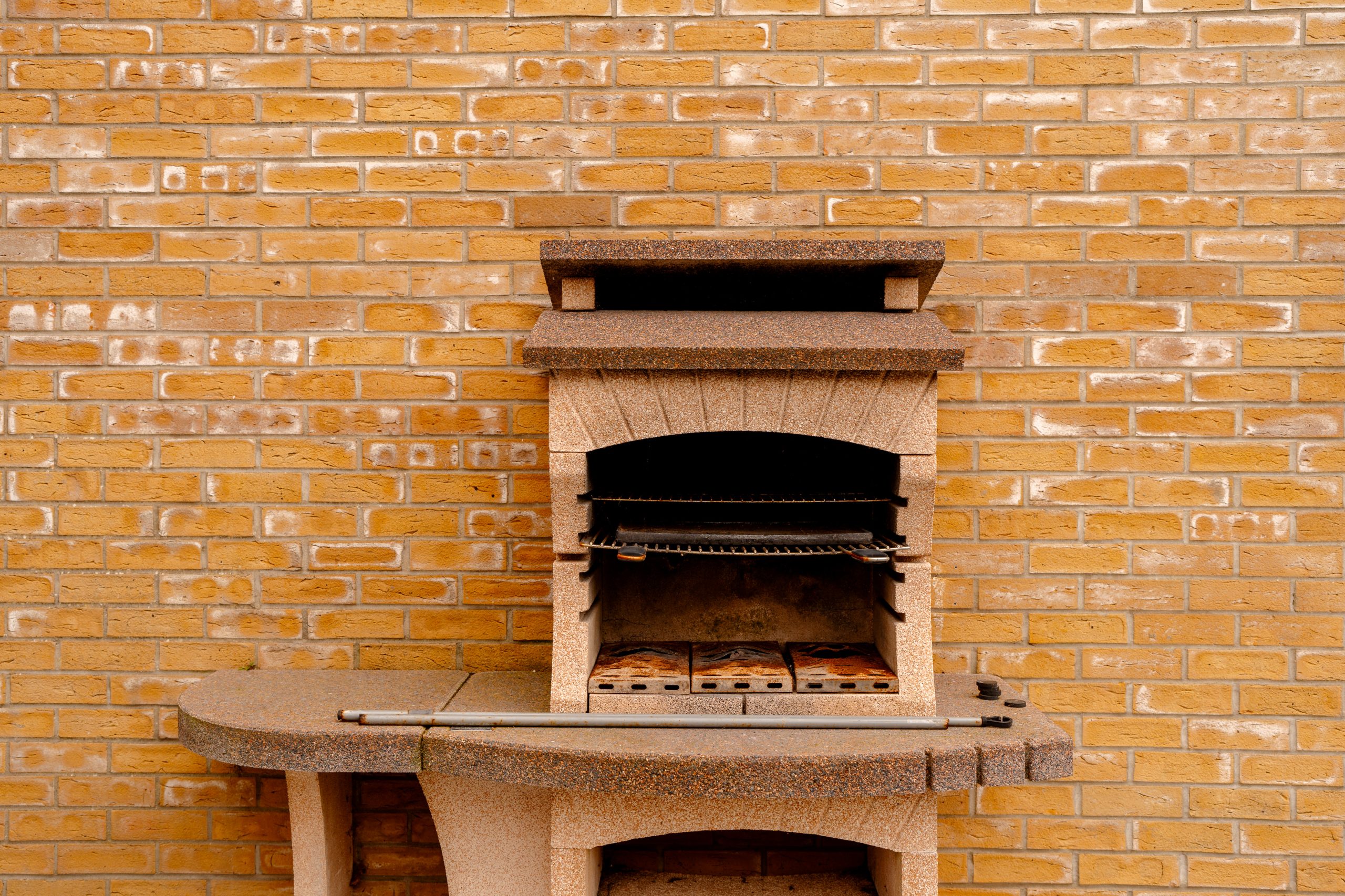 A stone outdoor barbecue grill with an oven sits against a yellow brick wall. The grill has a small chimney and a curved side table attached, providing space for food preparation or serving.