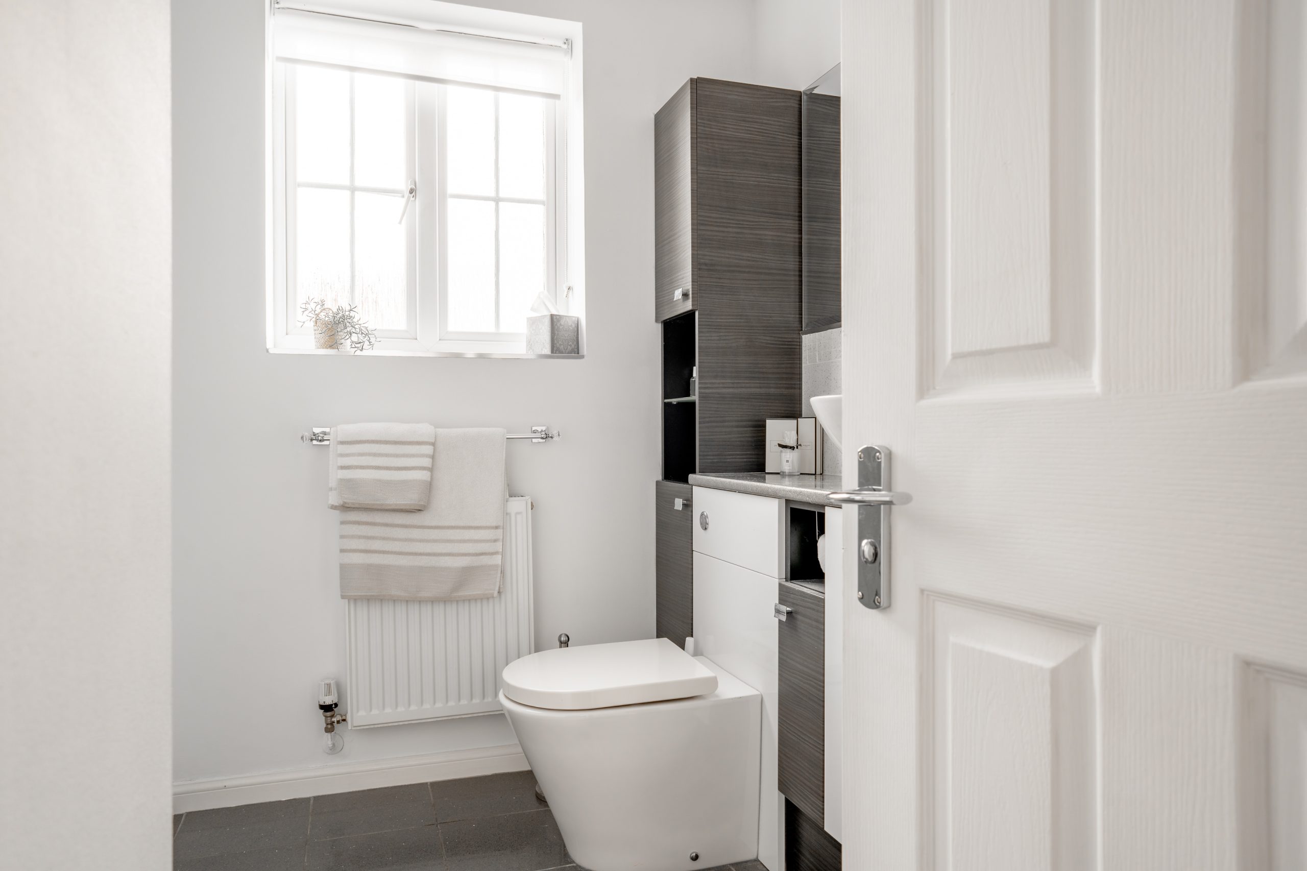 A modern bathroom with a closed white door, a toilet, and a dark wood cabinet. A white towel hangs on a radiator under a window with blinds. The floor is tiled in dark gray. Bright, natural light fills the space.