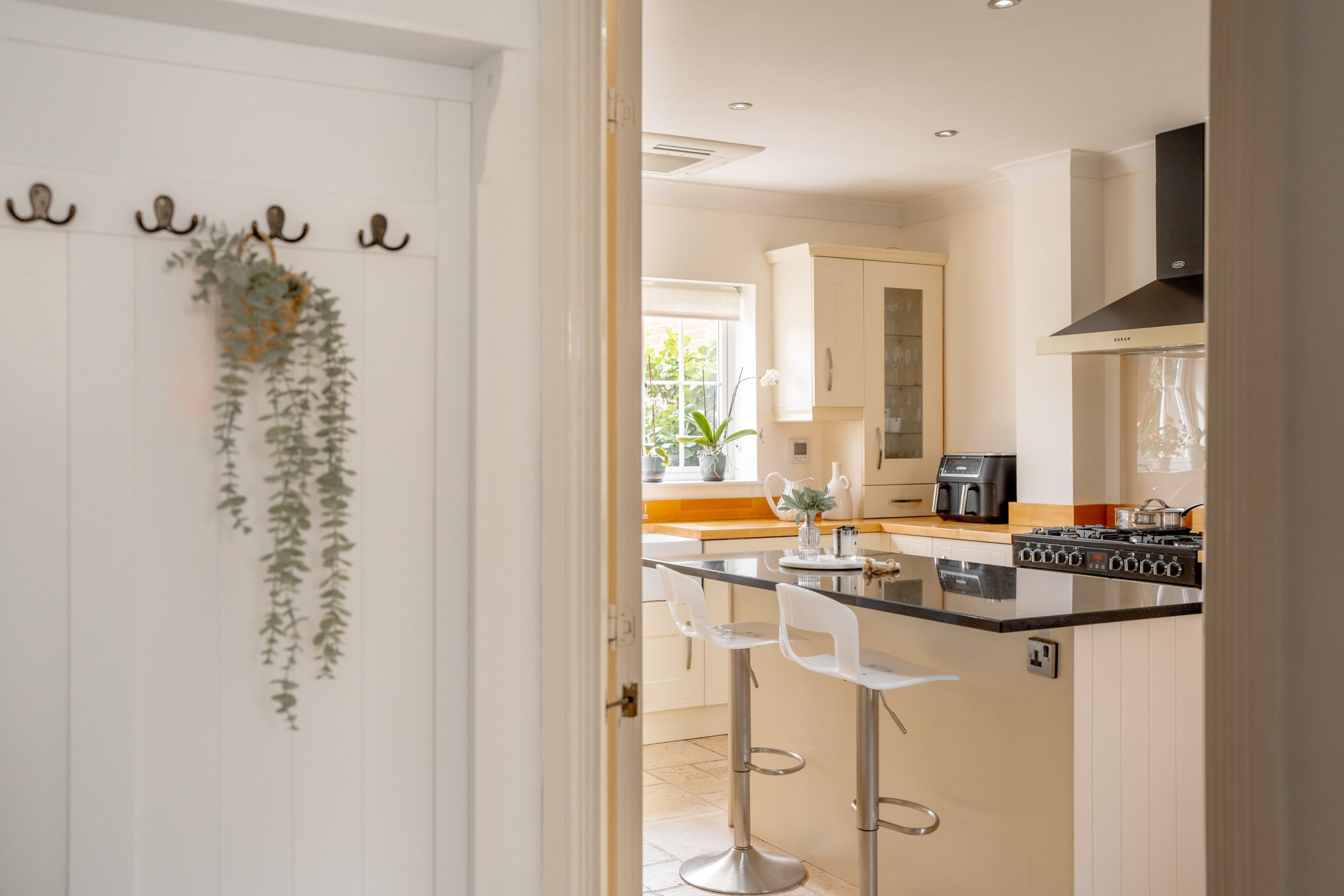 A modern kitchen with a central island, two bar stools, and stainless-steel appliances. A vase with flowers and a plant are on the countertop. The room is bright with natural light from a window. Cream cabinets and a black range hood are visible.