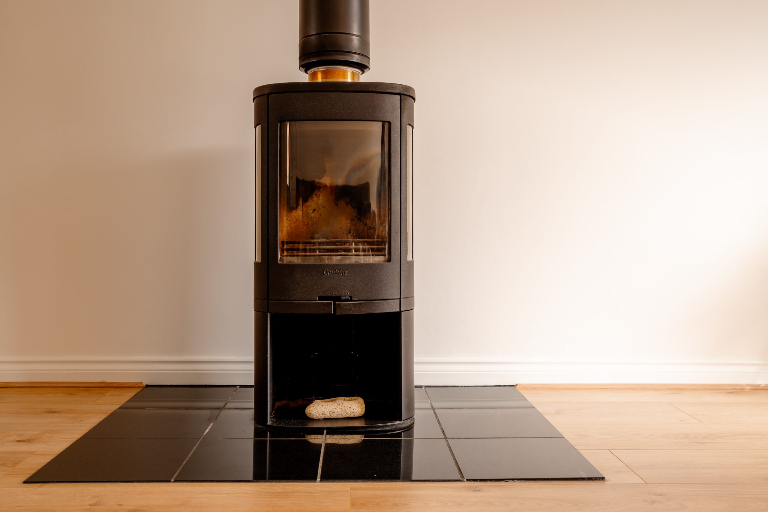 A black wood-burning stove on a tiled hearth against a beige wall, with a piece of firewood resting beneath it. The wooden floor contrasts with the sleek, modern design of the stove.