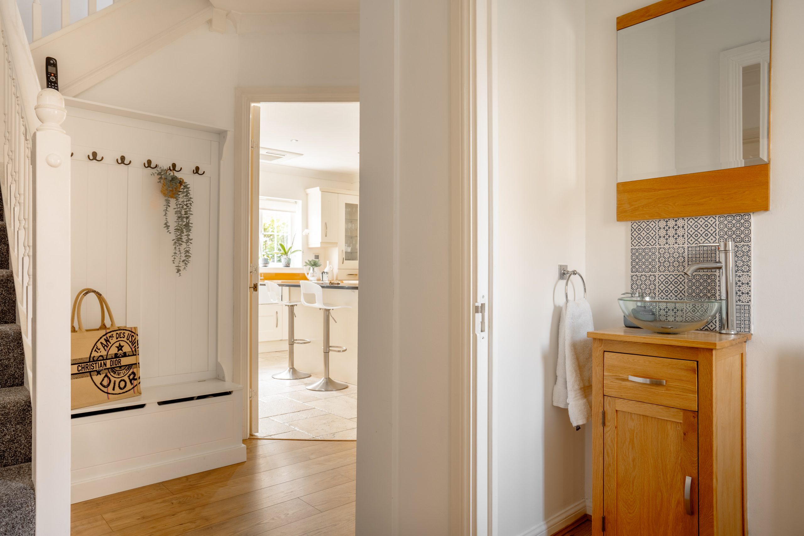 A bright, modern interior features a hallway with gray carpeted stairs, a white coat rack with hooks and storage, and a wooden cabinet with a glass sink and mirror. The hallway leads to a sunlit kitchen with white cabinets and bar stools.