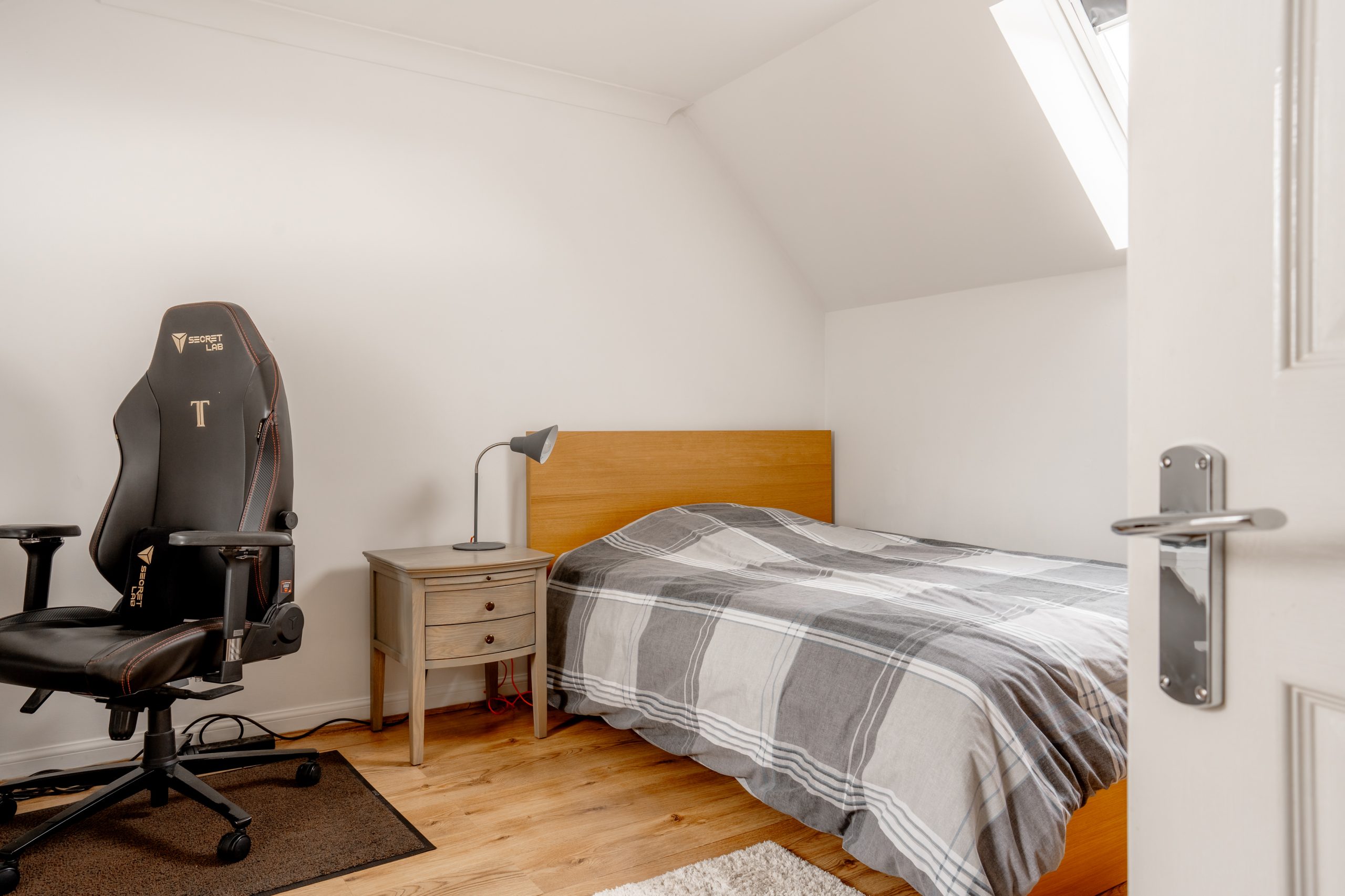 A cozy bedroom features a wooden bed with gray plaid bedding, a small nightstand with a lamp, and a black gaming chair on a rug. Natural light enters through a skylight, illuminating the wooden floor. A partially open door is visible.