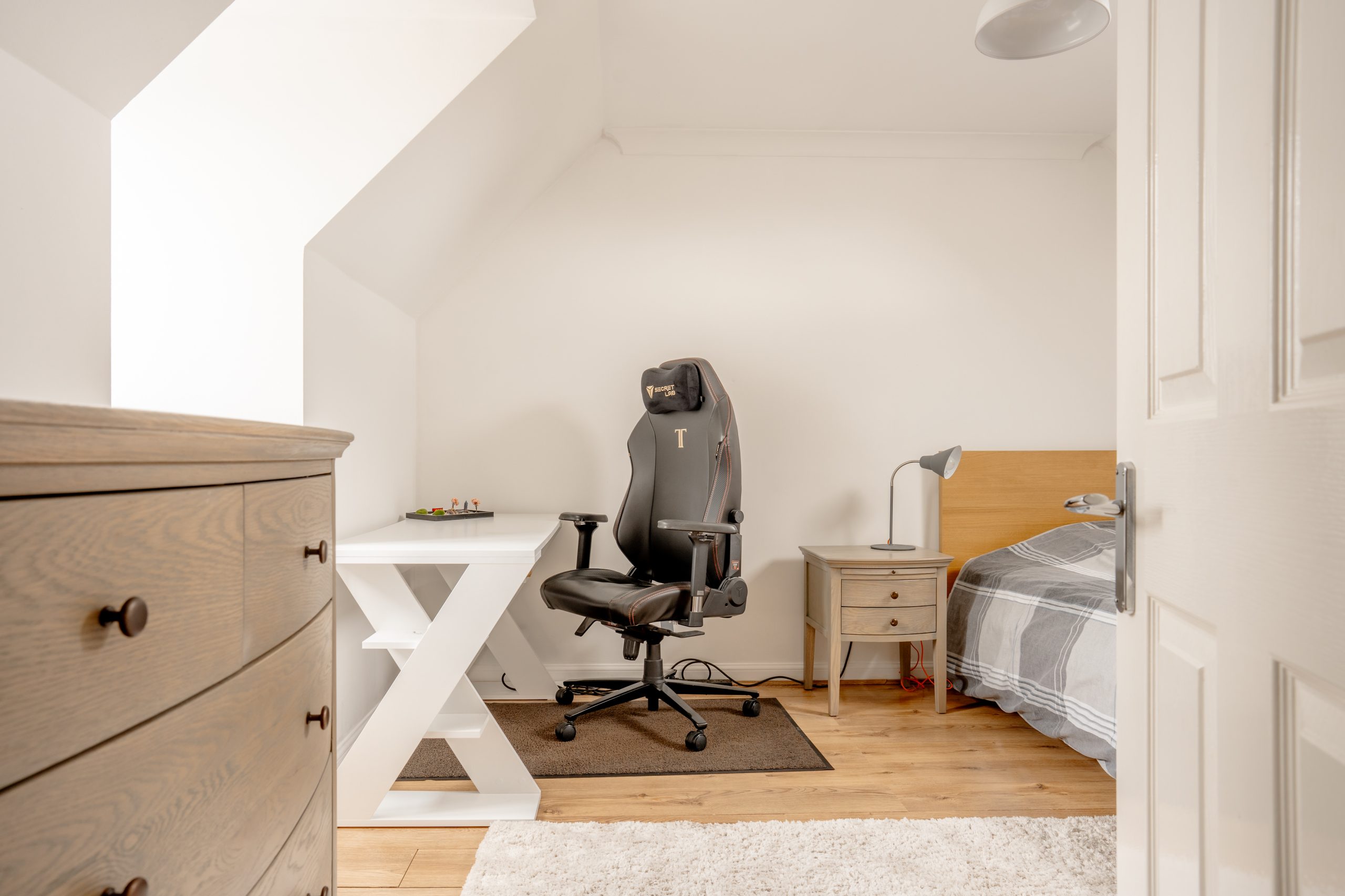 A cozy bedroom with a gaming chair at a white desk, a small lamp on a nightstand, and a neatly made bed with gray bedding. Light brown dresser and wooden flooring. Wall lamp above the desk. A white door is partially open, revealing the serene space.