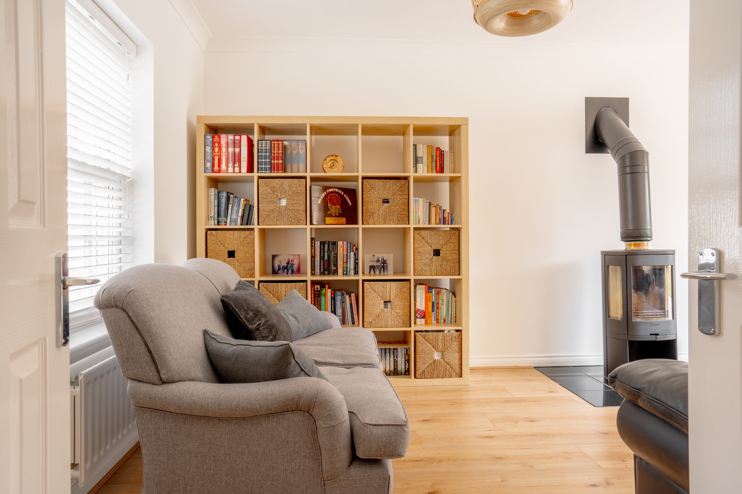 A cozy living room with a plush gray sofa, a wooden bookcase filled with books and baskets, and a modern black wood-burning stove on the right. The room has light wooden floors and white walls, with sunlight streaming through a window.