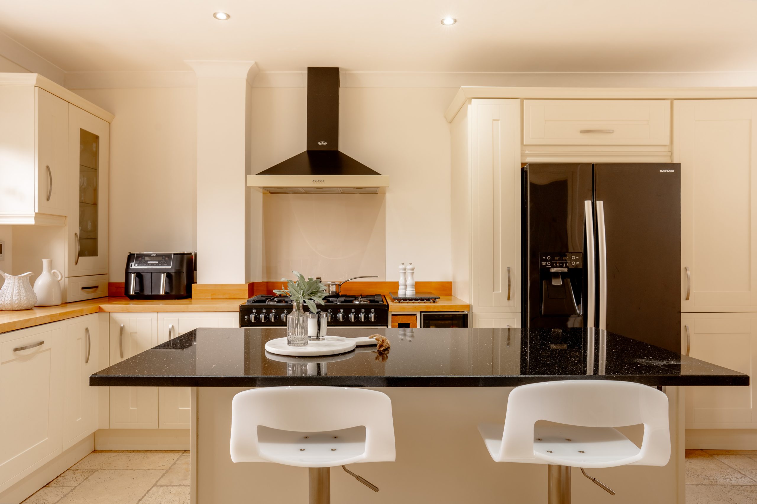 A modern kitchen with white cabinets, a black granite island, and two white barstools. It features a black refrigerator, a range cooker with a hood, and small appliances. A potted plant decorates the island countertop.