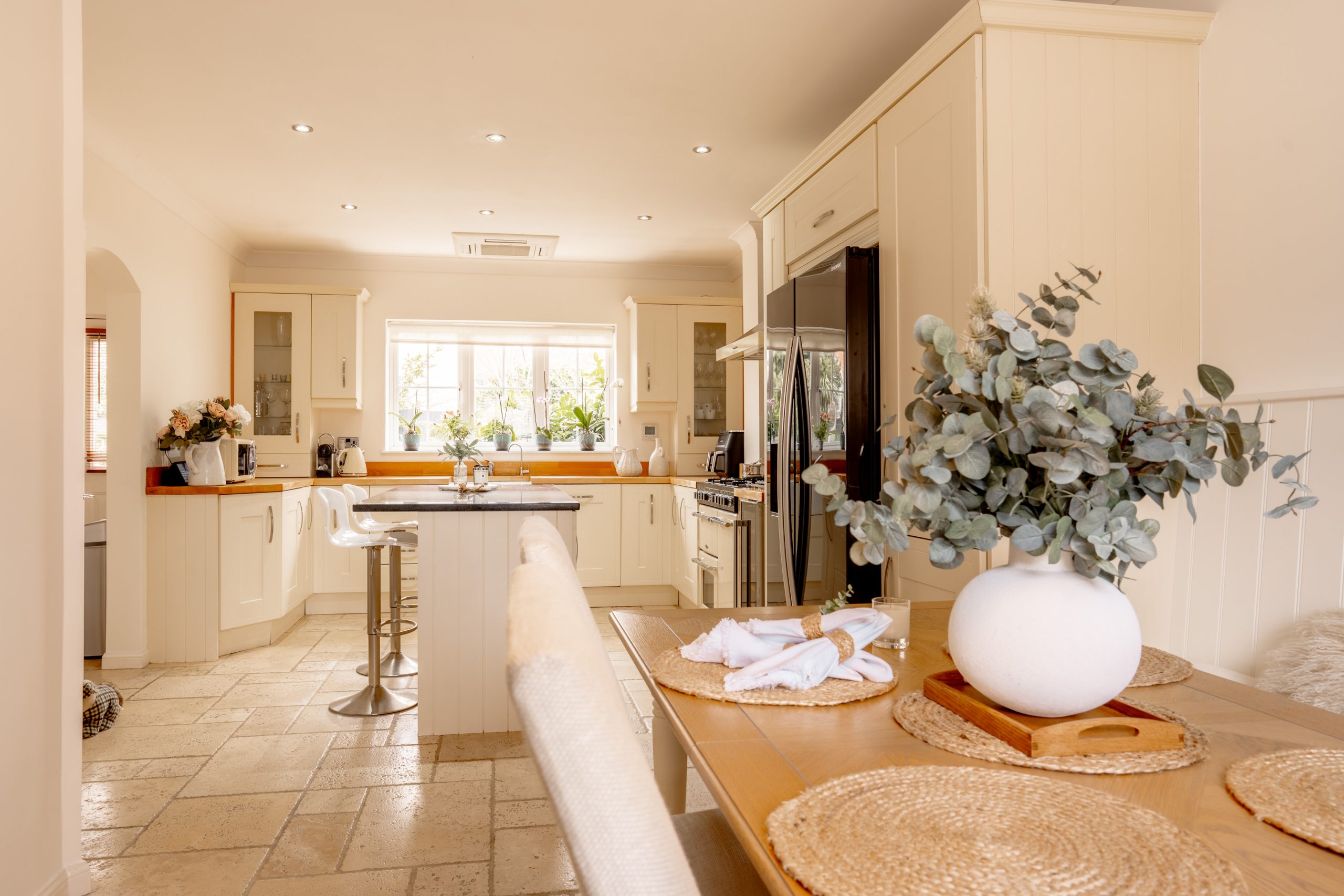 A bright kitchen with a central island and two bar stools. There's a black fridge, white cabinets, and a wide window letting in natural light. A dining table with a vase of eucalyptus branches and woven placemats is in the foreground.