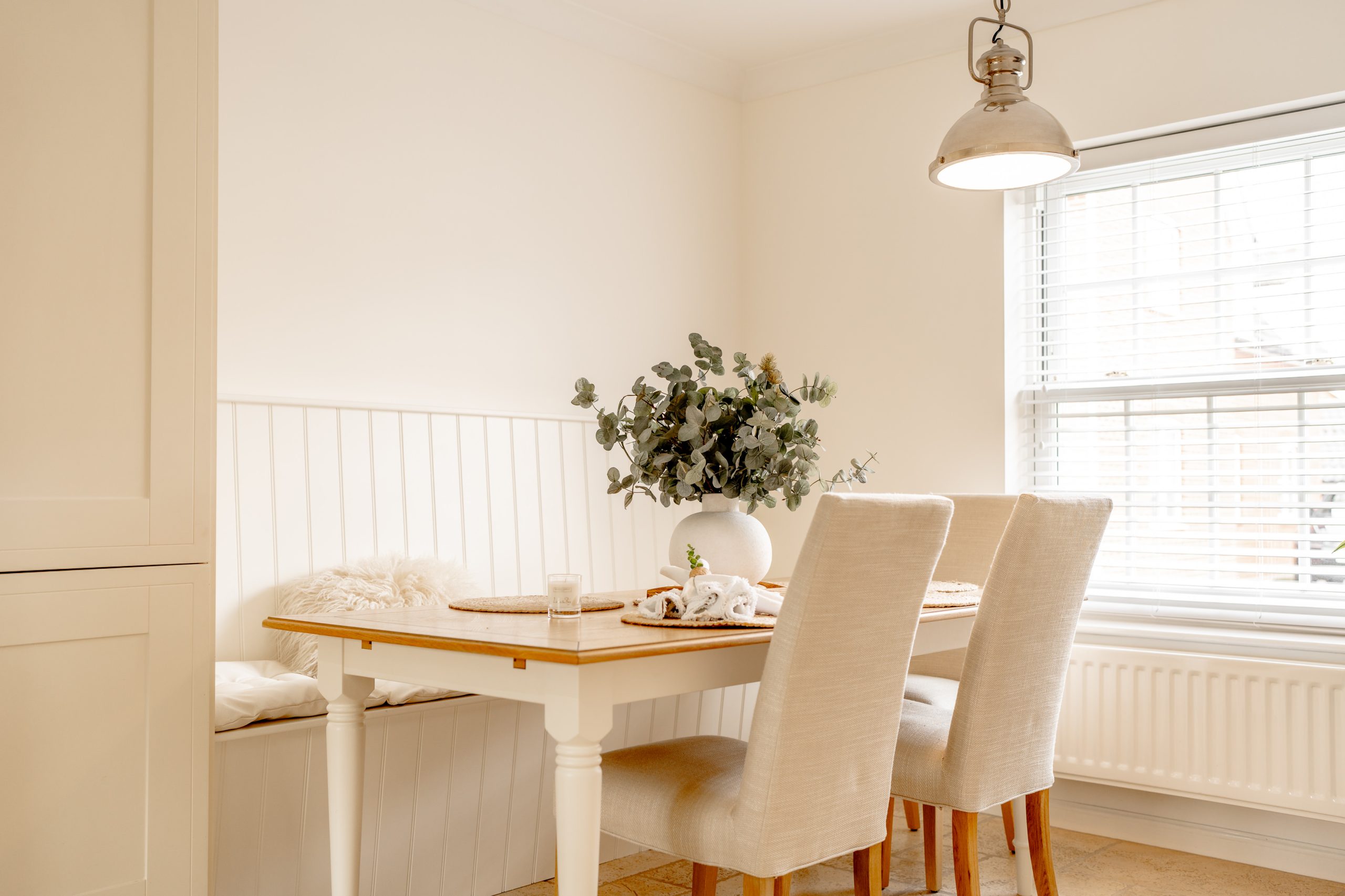 A cozy dining area with a wooden table, two beige chairs, and a bench. A vase with green foliage is centered on the table. Natural light streams in through a window with white blinds. A pendant lamp hangs above, and a cushion rests on the bench.