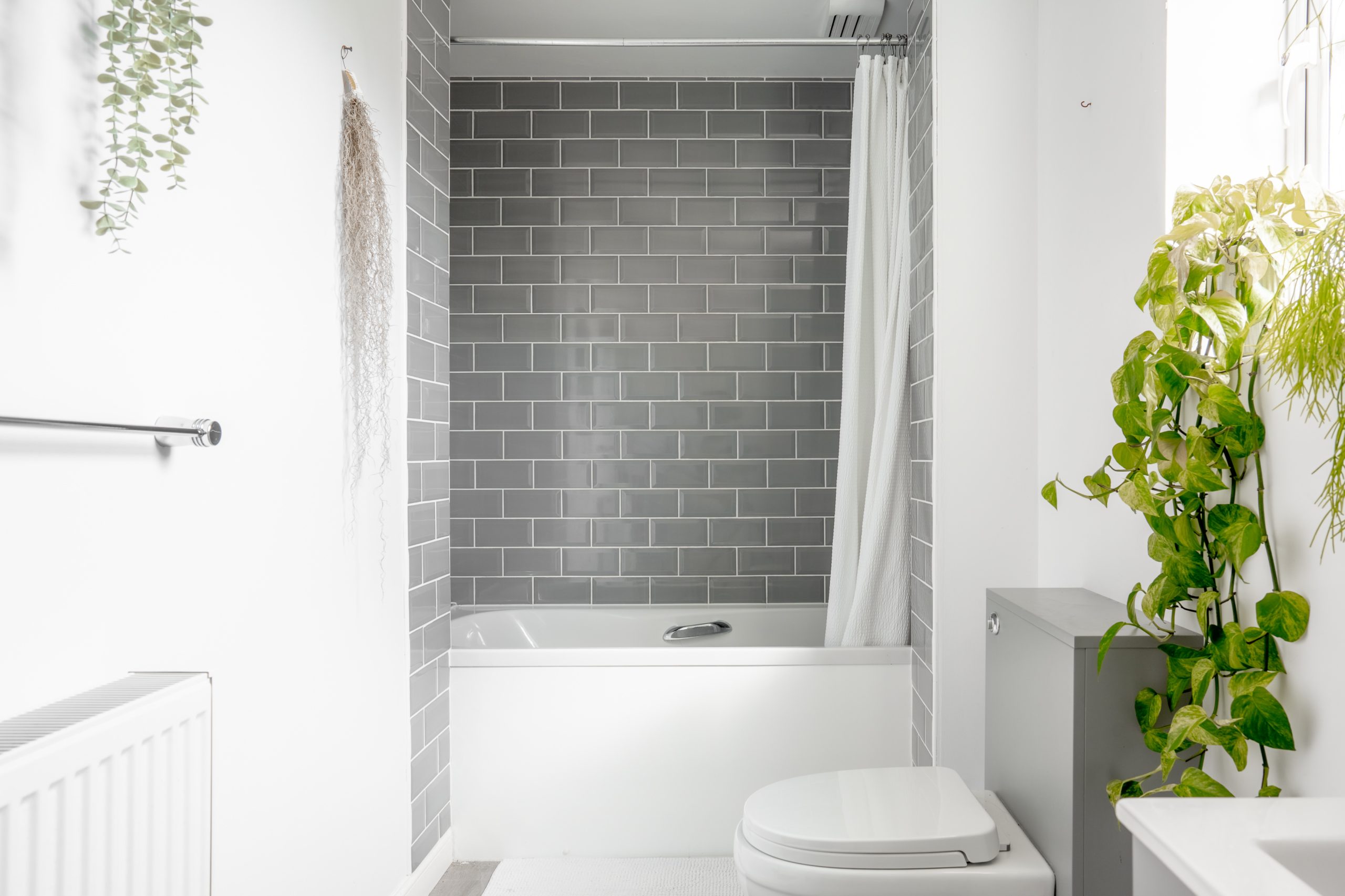 A modern bathroom with gray subway tiles in the bathtub area, a white shower curtain, a toilet, and lush green plants on the right. Bright white walls and a radiator are visible on the left.