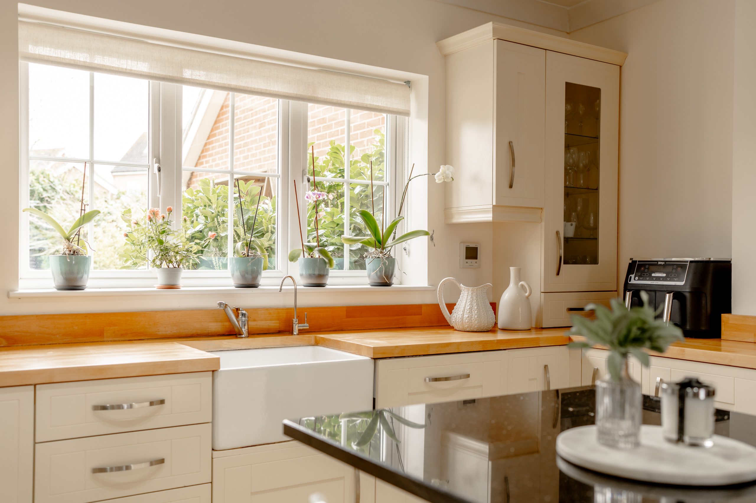 A bright kitchen with white cabinets and wooden countertops. There's a farmhouse sink beneath a window with potted plants on the sill. A black countertop island in the foreground has a plant and seasonings. A coffee maker and decor are on the corner counter.
