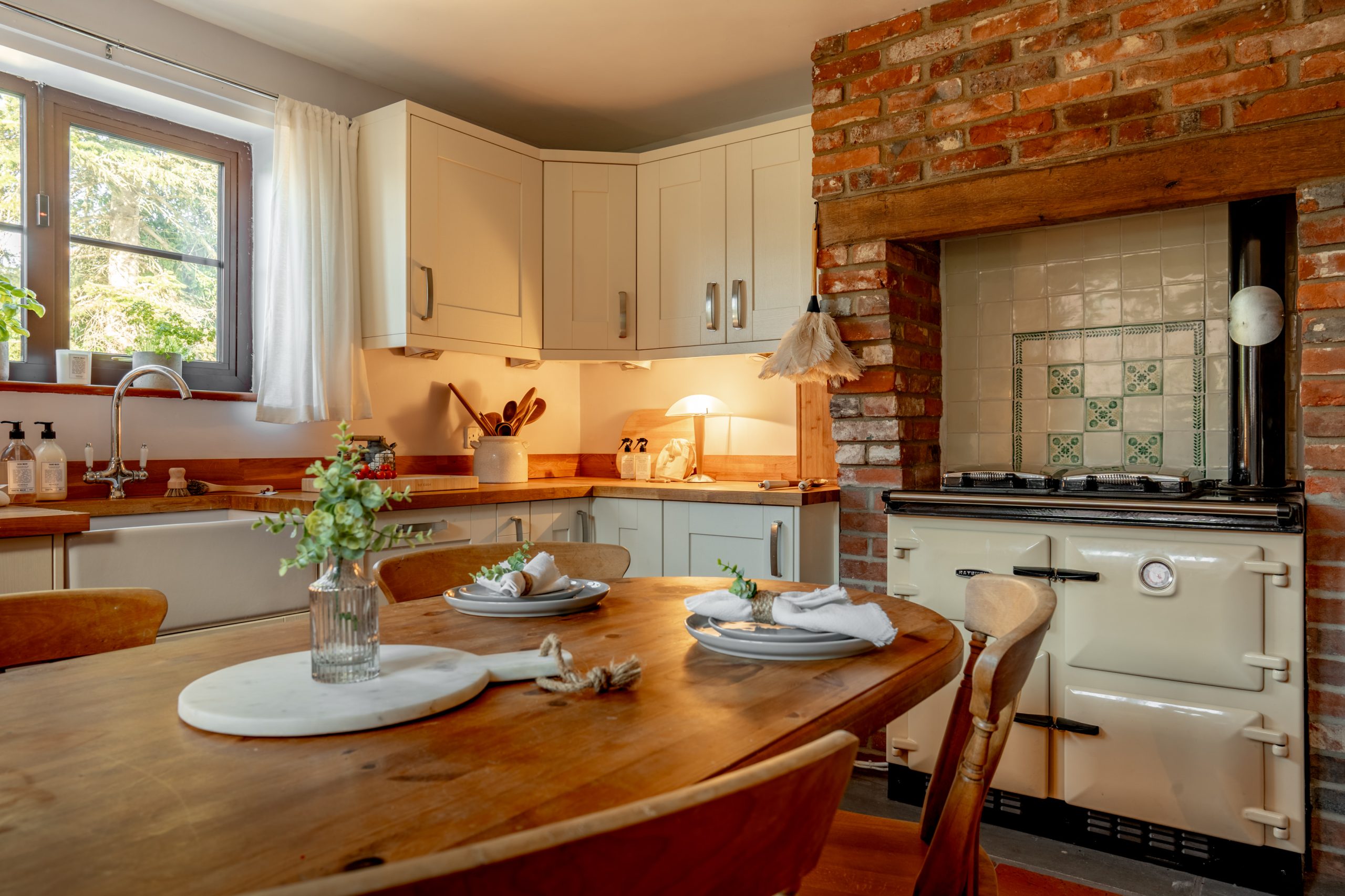 A cozy kitchen with a rustic design features a wooden dining table set for four. White cabinets and a brick accent wall surround a vintage cream stove. A vase with flowers sits on the table, and a window provides natural light.