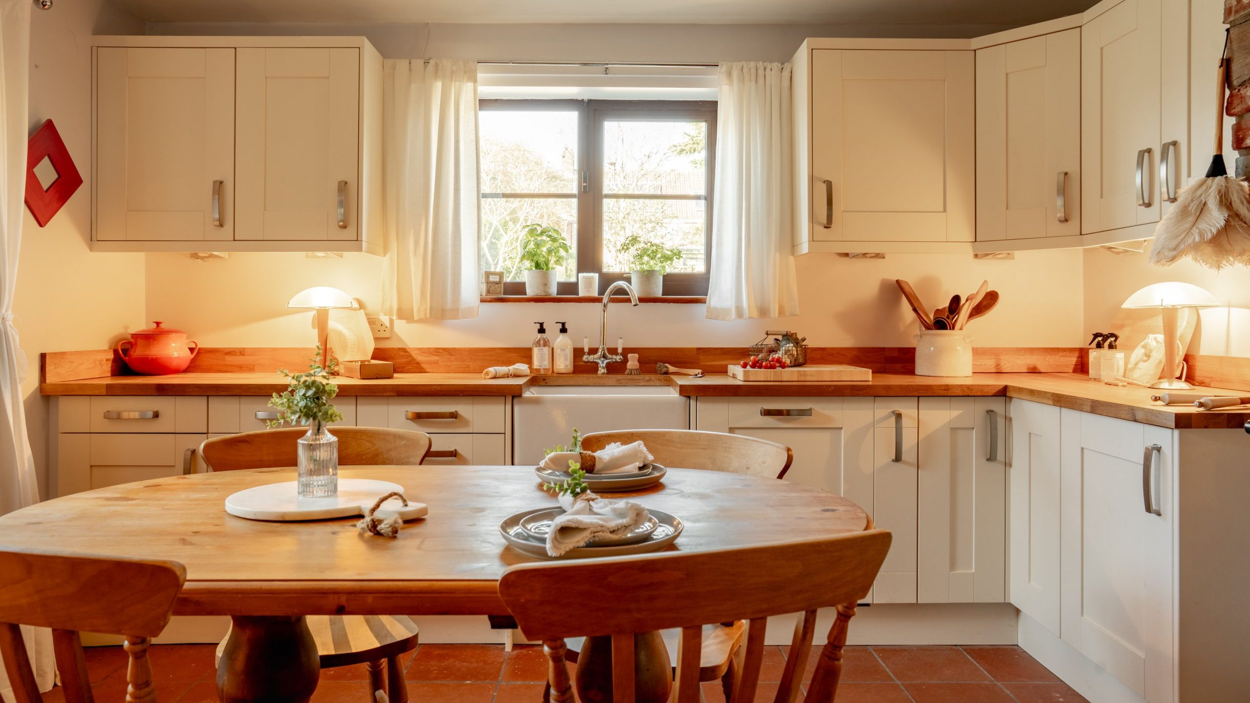 A cozy kitchen with wooden furniture, white cabinets, and a wooden dining table set with plates. Soft lighting creates a warm atmosphere. A window with curtains provides natural light, highlighting the red teapot on the counter.