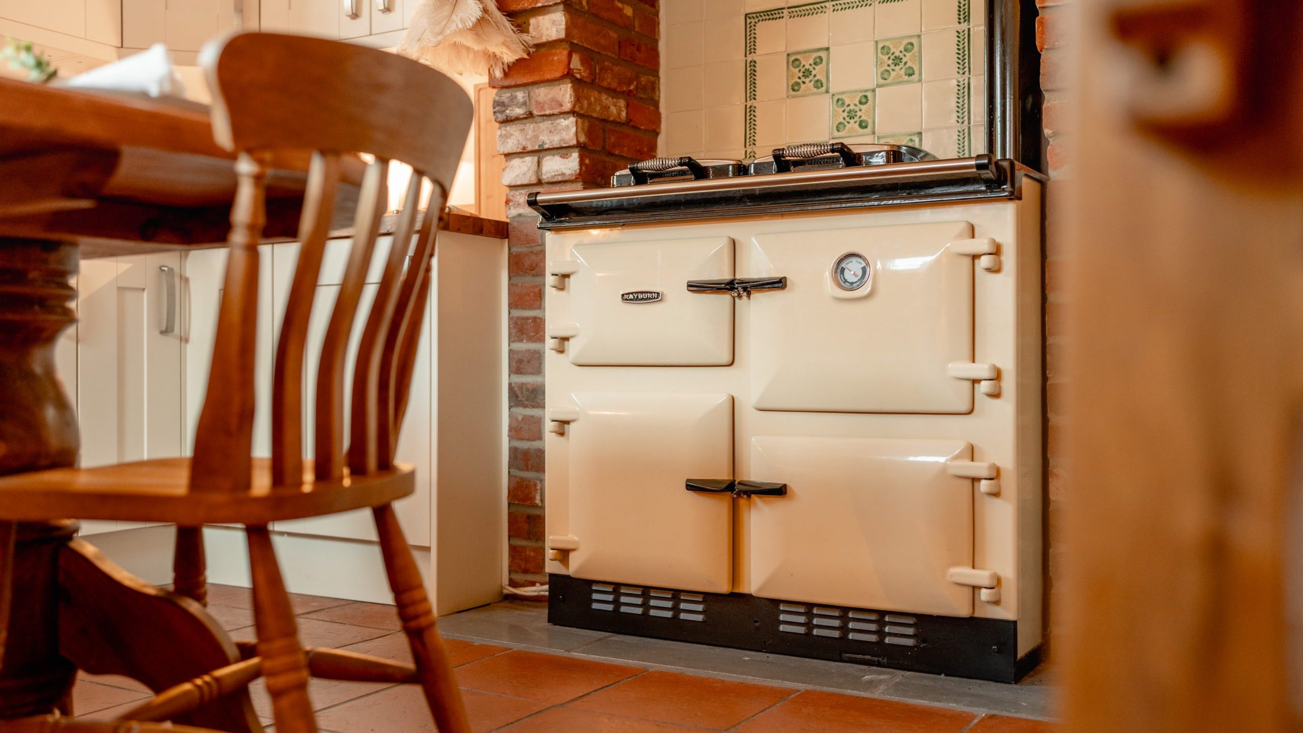 A cream-colored vintage stove with four compartments stands against a brick wall, next to white kitchen cabinets. A wooden chair and dining table are partially visible on the left. The floor is made of terracotta tiles.