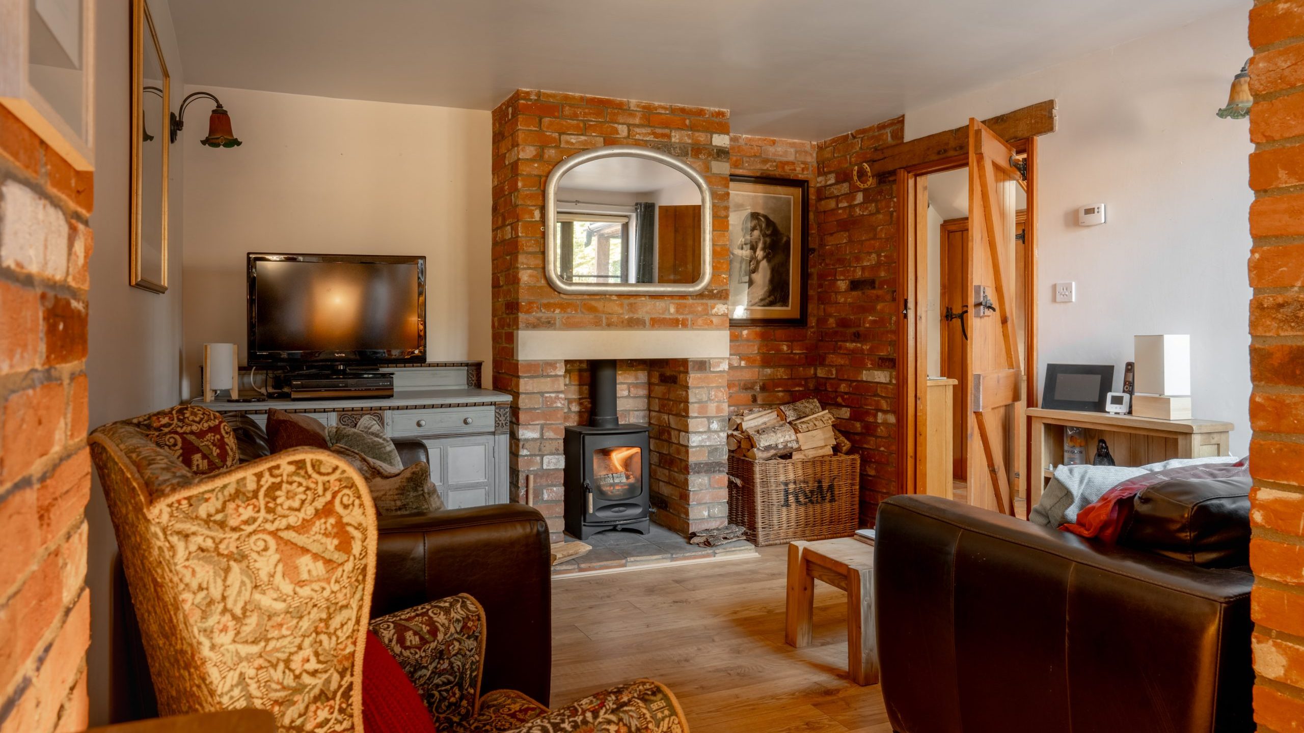 A cozy living room with brick walls features a wood-burning stove, TV, and armchairs. A framed portrait and mirror hang above the mantle. A wooden door and basket are adjacent to a console table with a speaker. Natural light fills the space.