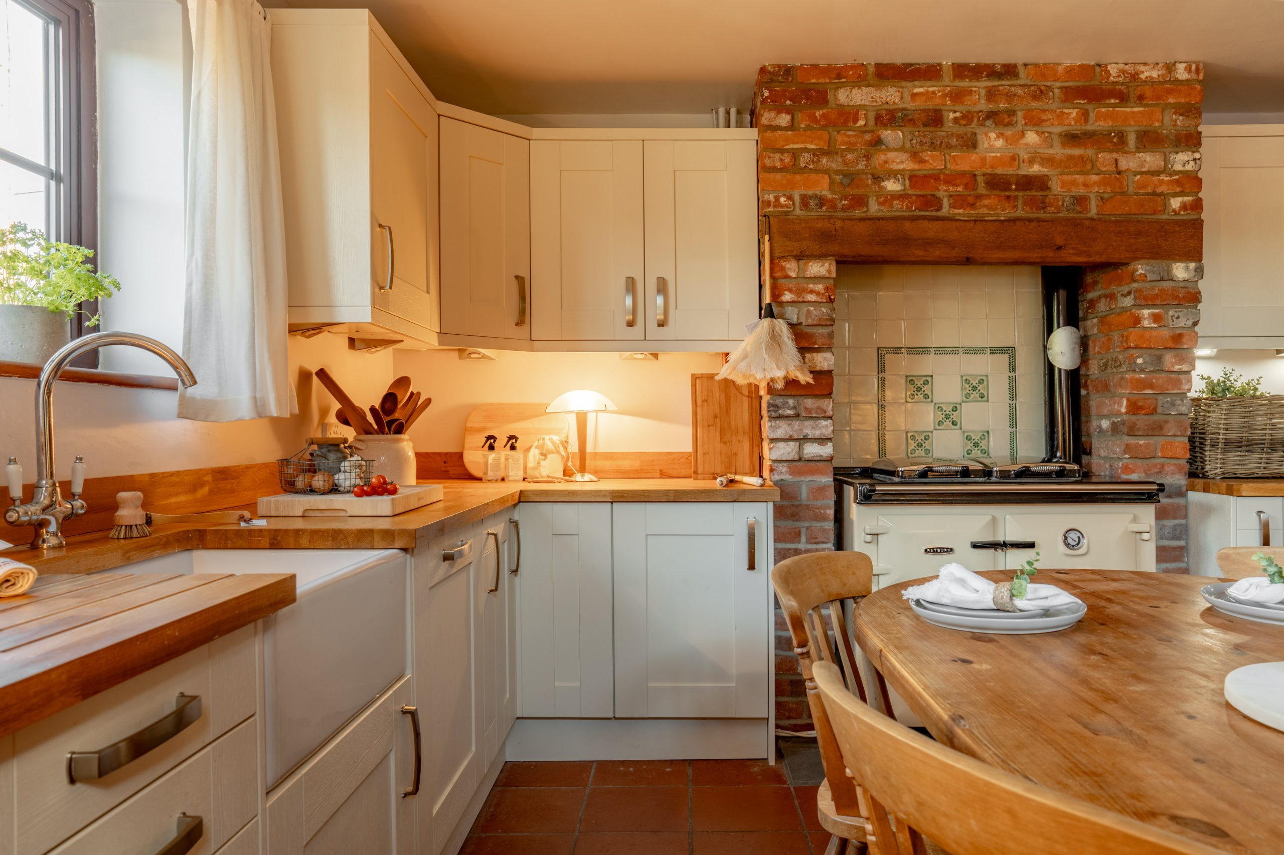A cozy kitchen with wooden countertops and white cabinets. An old brick arch surrounds a vintage stove. A round wooden table with chairs is set with plates. Natural light streams in through a window with a small plant on the sill.