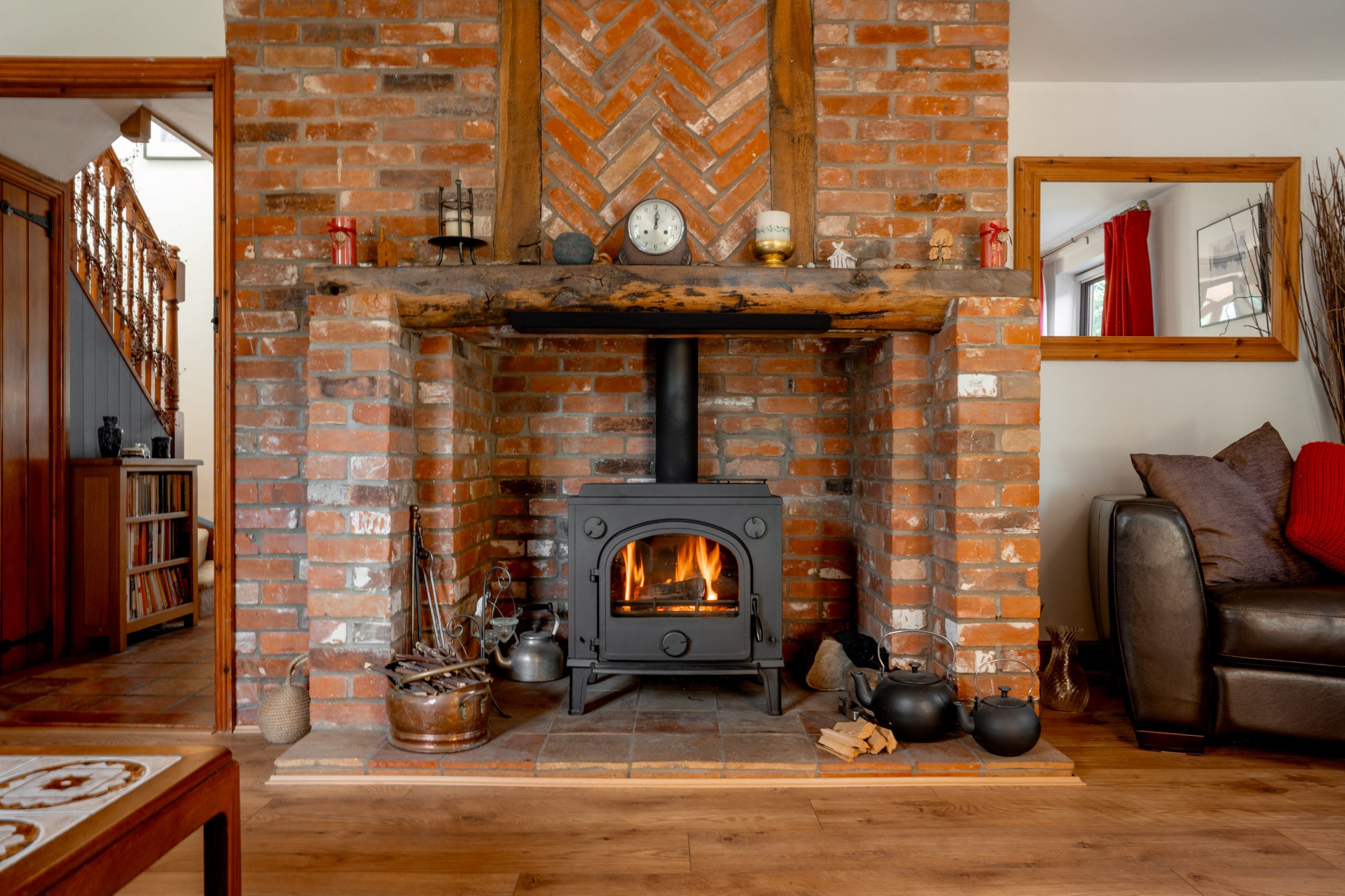 A cozy living room features a wood-burning stove set in a brick fireplace. The fire is lit, creating a warm glow. The rustic decor includes a wooden mantle, pots, and a basket of logs. A couch with red cushions sits to the right.