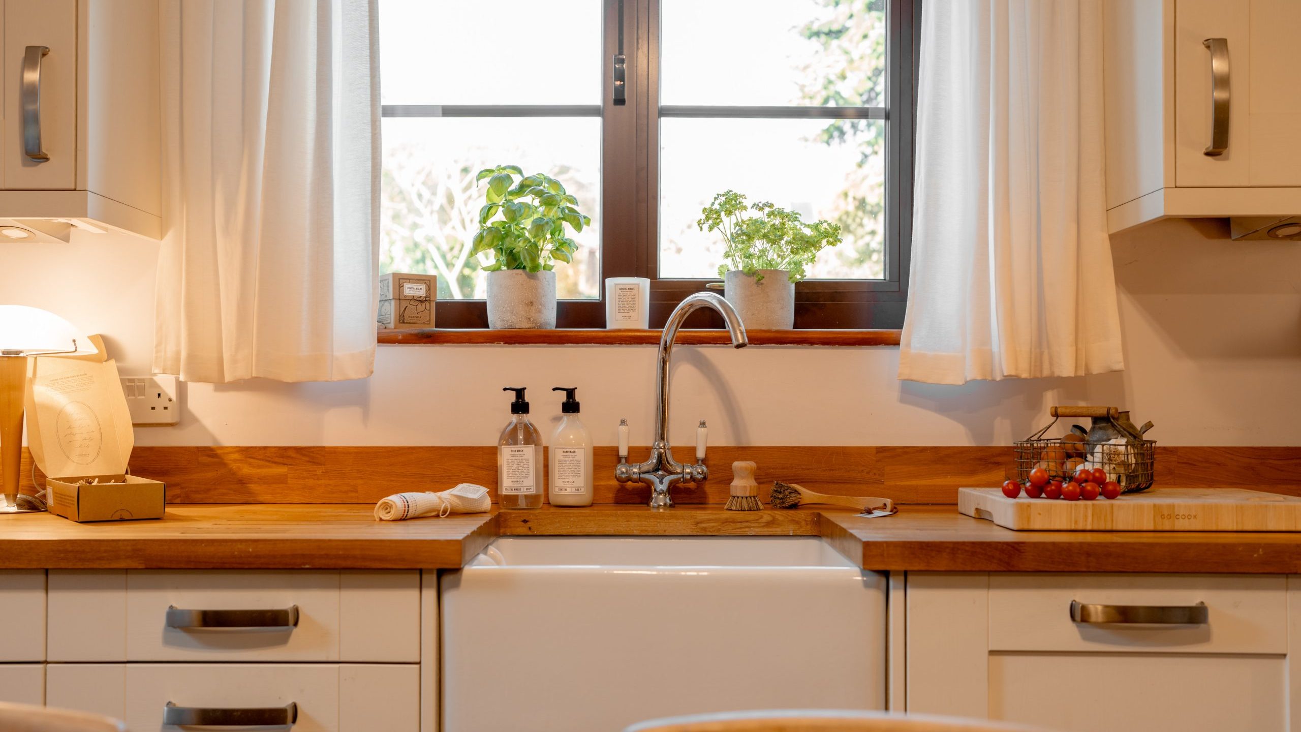 A cozy kitchen with a farmhouse sink, wooden countertops, and white cabinets. Two potted plants sit on the window sill above the sink. Soap dispensers and a towel are nearby, and tomatoes are on a cutting board to the right.