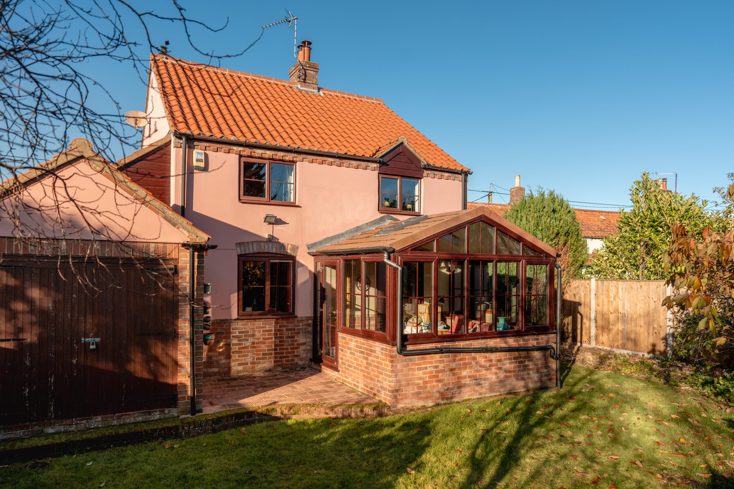A cozy two-story house with a red-tiled roof and pink exterior. It features a glass conservatory attached to the side, surrounded by a lush green lawn. The clear blue sky and late afternoon sunlight enhance the warm, inviting atmosphere.