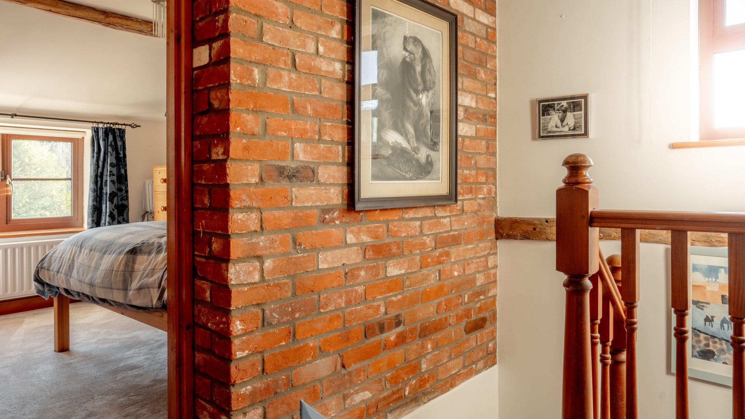 A cozy bedroom hallway with a rustic brick wall featuring framed black-and-white artwork. Wooden railings lead to a staircase, and a window lets in light, illuminating the bed and curtains in the adjacent room.