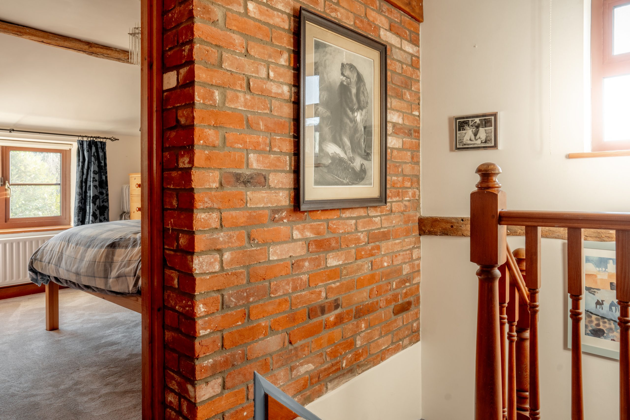 A cozy bedroom hallway with a rustic brick wall featuring framed black-and-white artwork. Wooden railings lead to a staircase, and a window lets in light, illuminating the bed and curtains in the adjacent room.