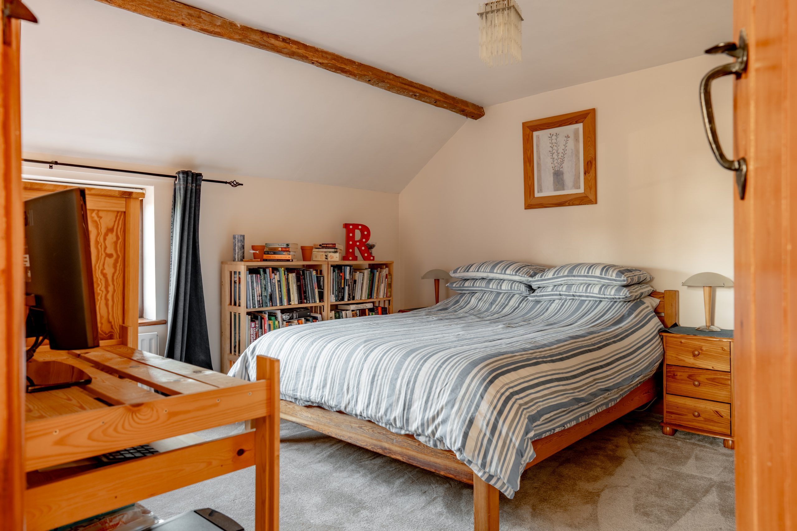 A cozy bedroom with a striped duvet-covered bed, wooden beams on the ceiling, and a framed picture on the wall. There's a bookshelf filled with books and decor, a small desk with a monitor, and a window with gray curtains letting in natural light.
