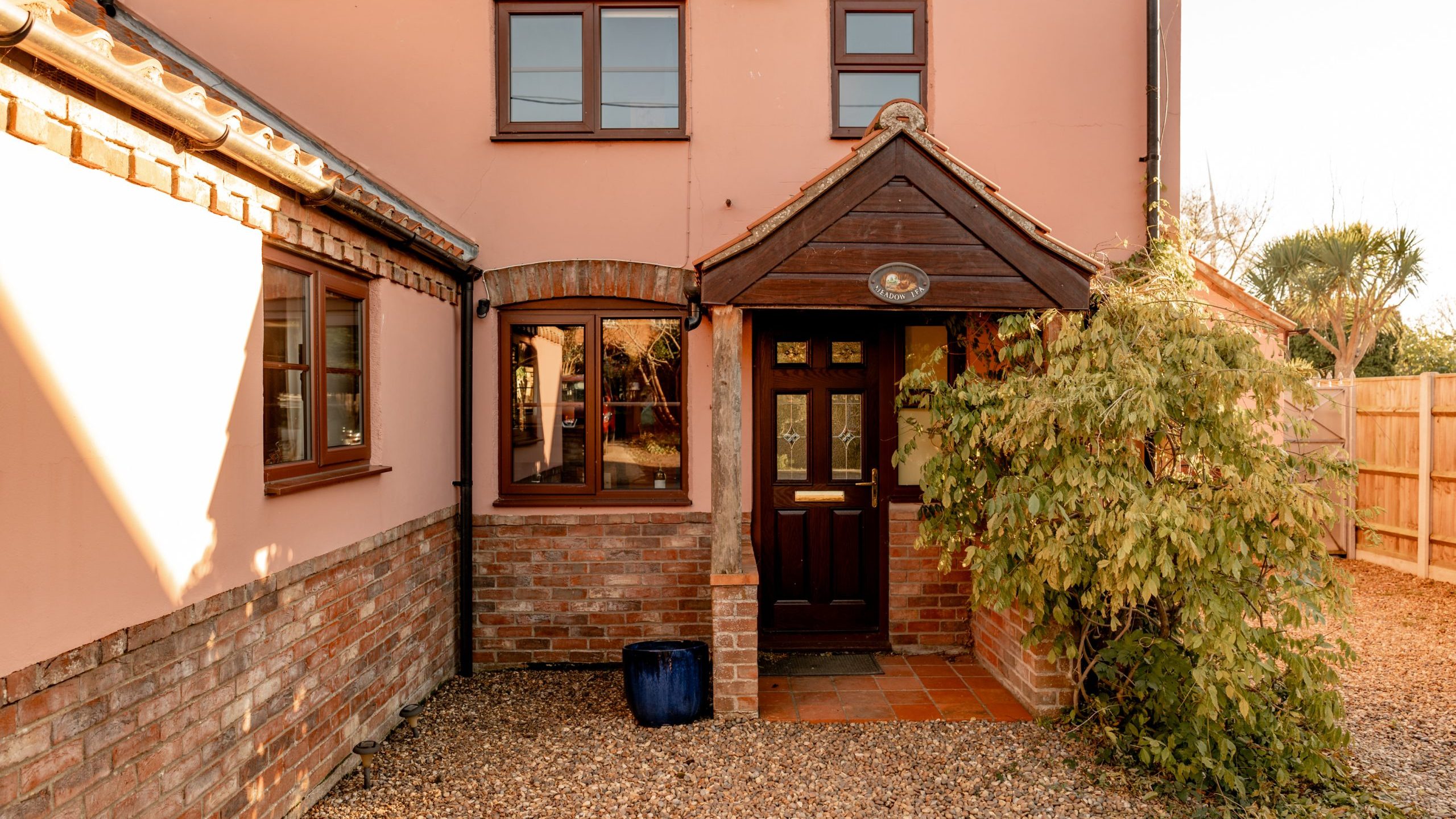 A charming two-story house with a pink exterior and brick accents. It has large windows, a dark wooden door, and a small porch with plants. The front yard is covered with gravel, and a wooden fence is visible on the right side.