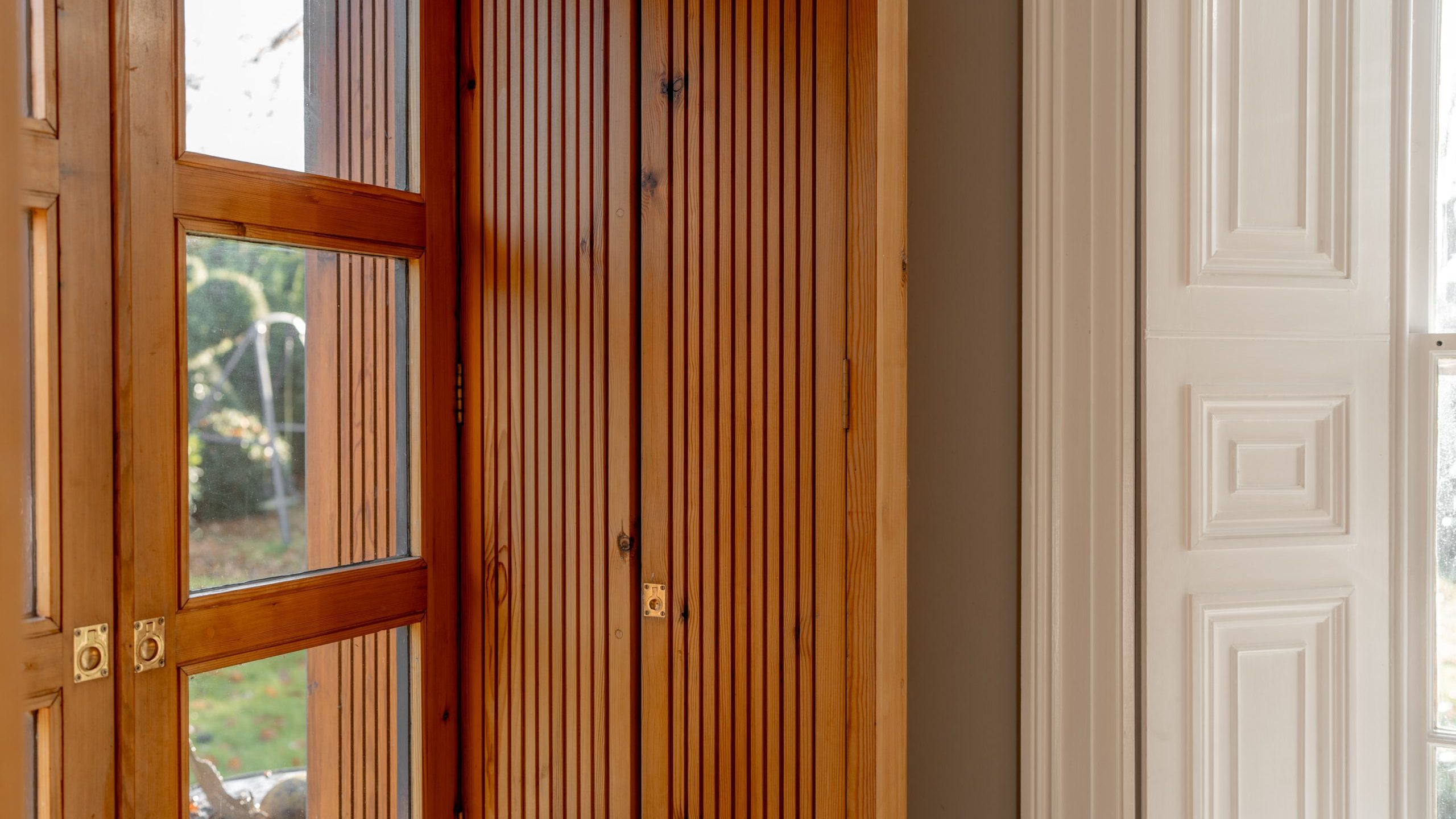 A close-up of a wooden door with slatted panels and glass sections, adjacent to a white paneled wall. Sunlight softly illuminates the area, casting gentle shadows. Outside, foliage and a blurred background are visible through the glass.