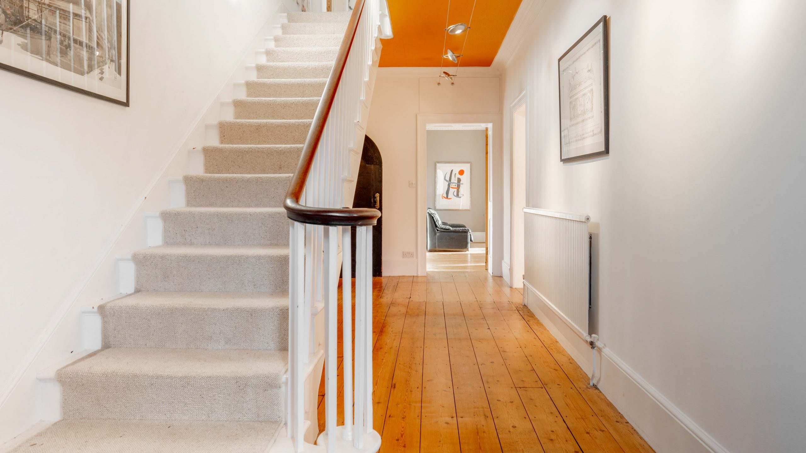 A hallway with wooden floors and white walls features a carpeted staircase on the left. Overhead, there are track lights on a bright orange ceiling. Framed pictures adorn the walls, and a room with a chair is visible at the end of the hallway.