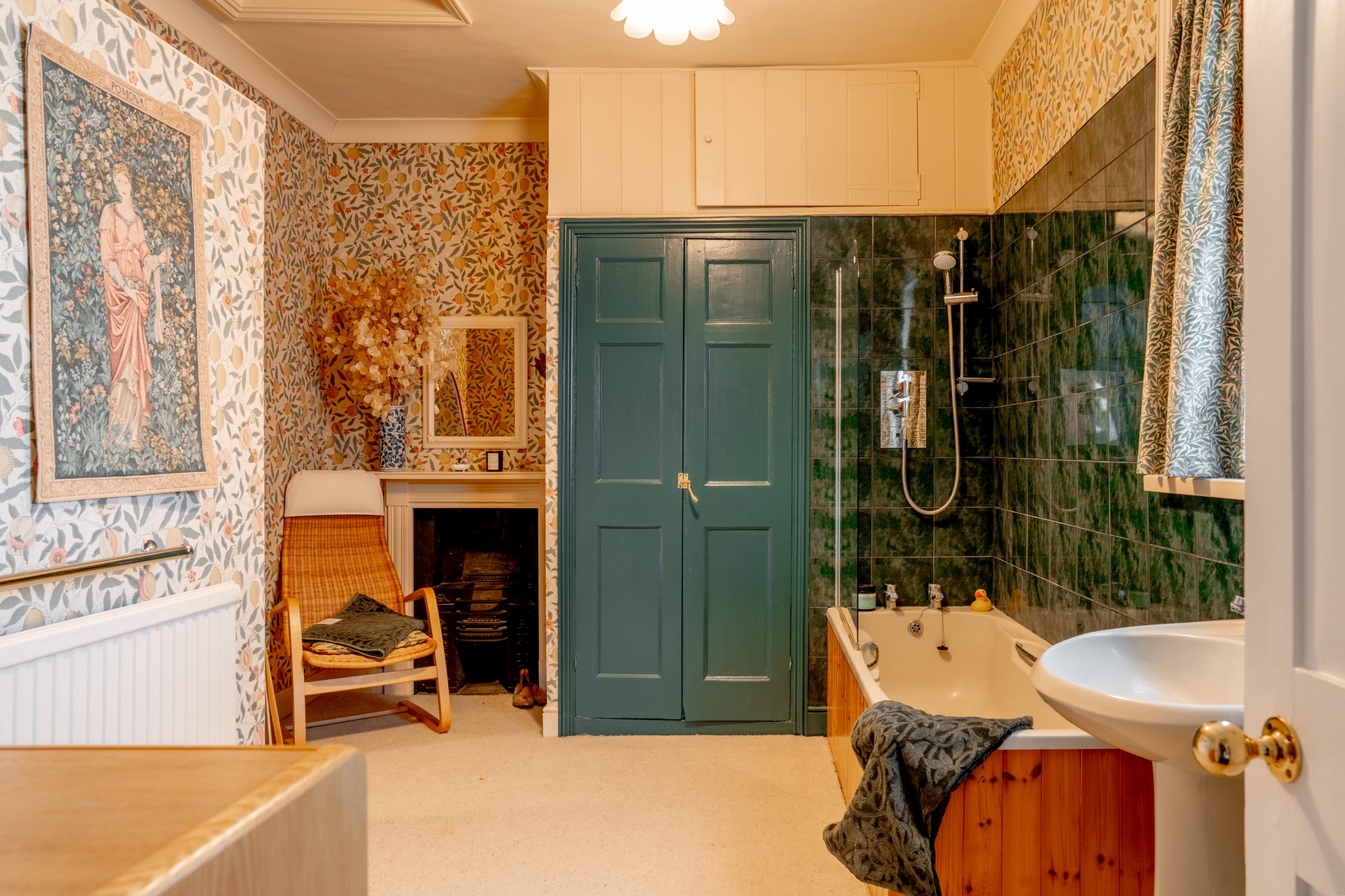 A cozy bathroom with floral wallpaper and dark green tiles. It features a bathtub with a shower, a white sink, and a large green door. Decorative art and a wicker chair add a touch of warmth and elegance.