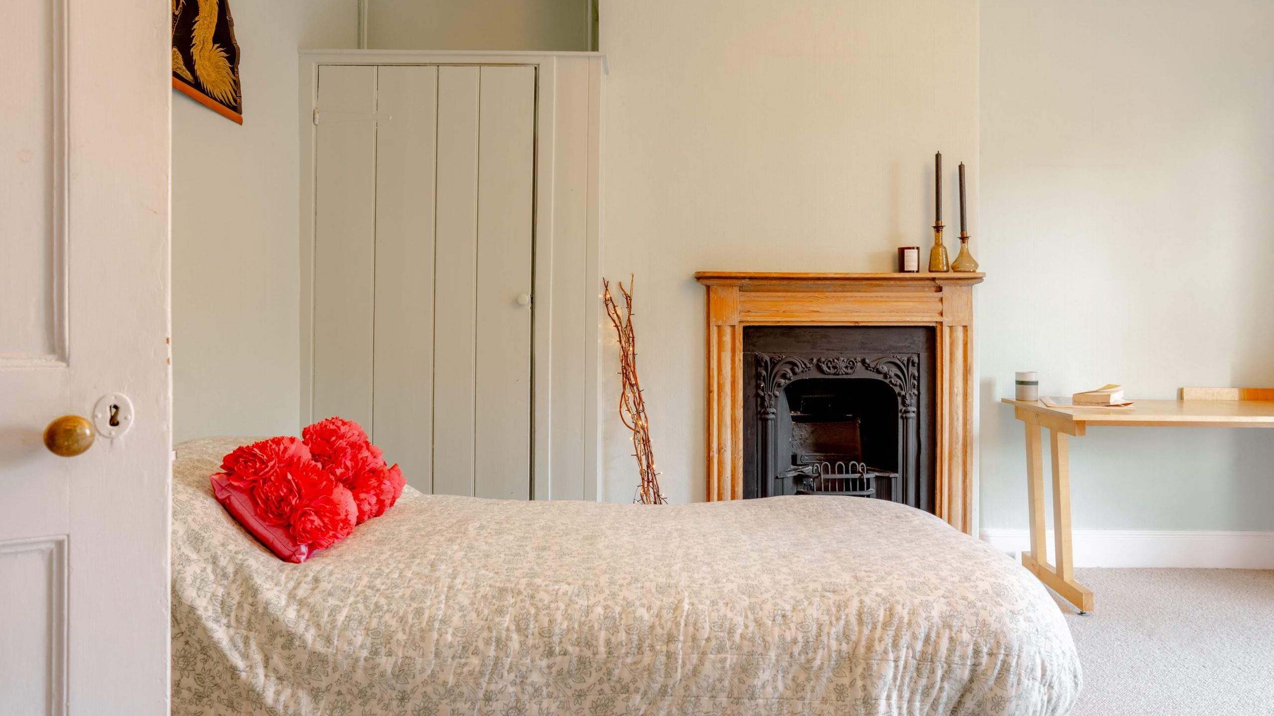 A cozy bedroom with a light green wall, featuring a bed with floral bedding and red cushions. There's a wooden fireplace with decorative candles, a minimalist desk, and a closed door leading to a closet.