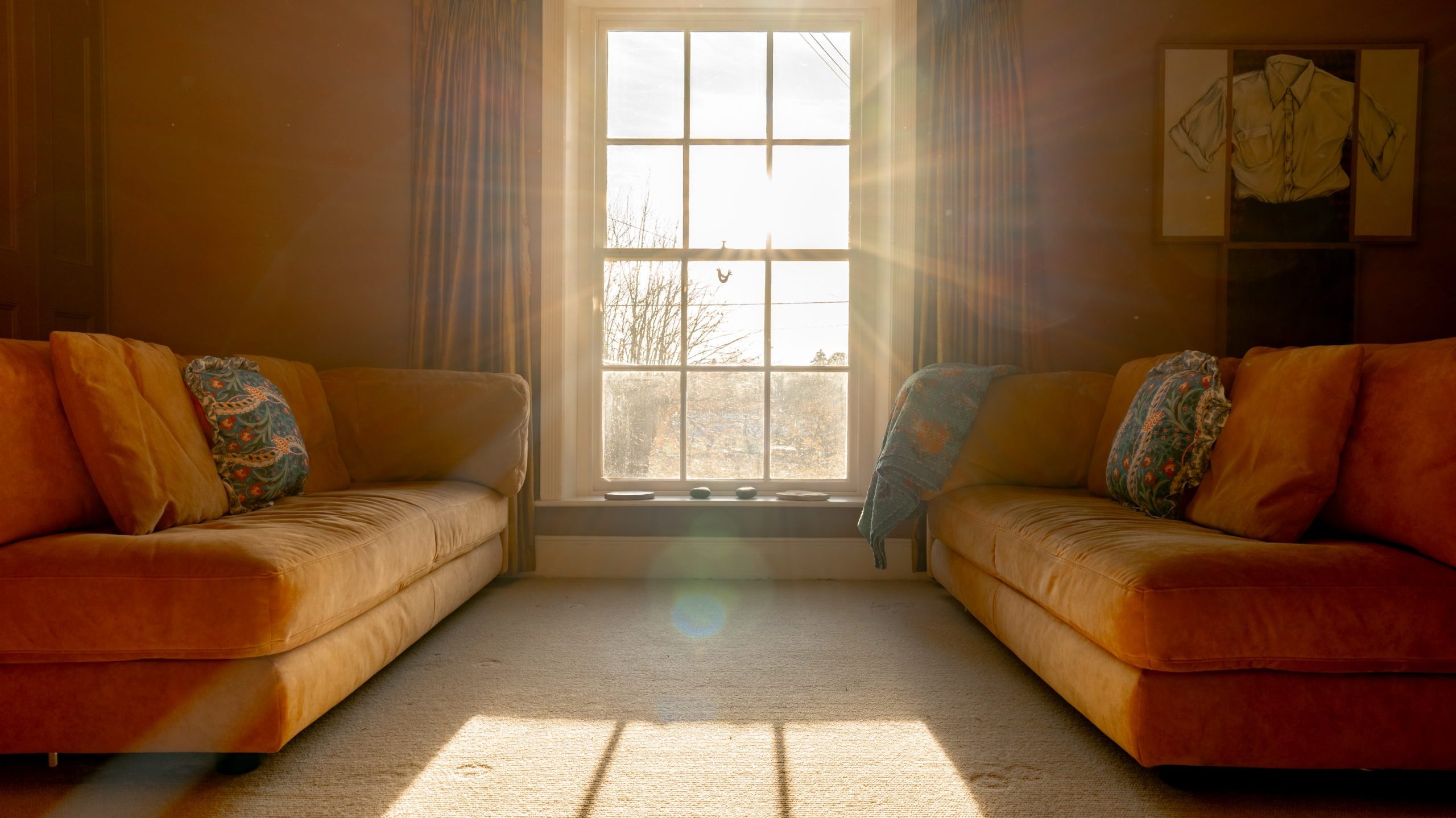 Sunlit living room with two orange sofas facing each other, featuring colorful cushions. A large window between the sofas lets in bright sunlight, casting long shadows on the carpeted floor. Framed shirts hang on the wall beside the window.