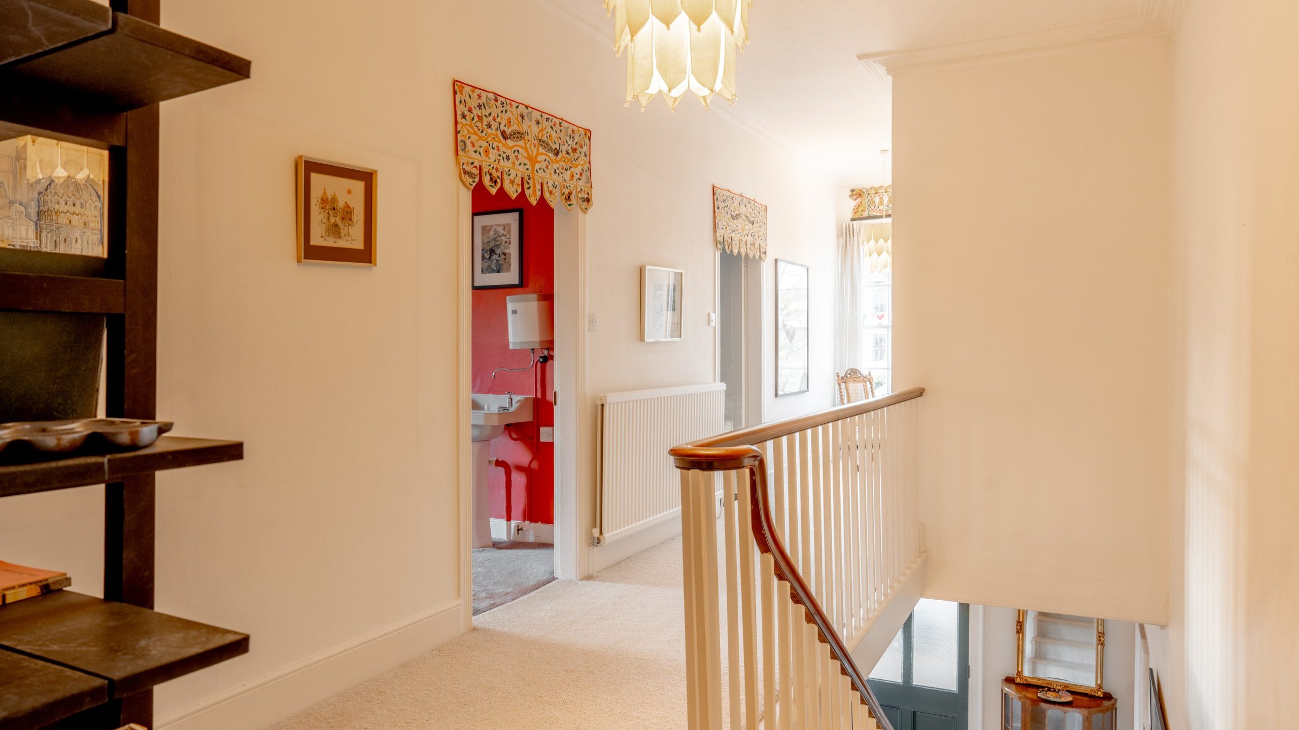 A bright hallway with cream walls and carpet, featuring a wooden railing on the staircase. A shelf with decorative items is on the left, and a doorway leads to a room with red walls. A window lets in natural light.