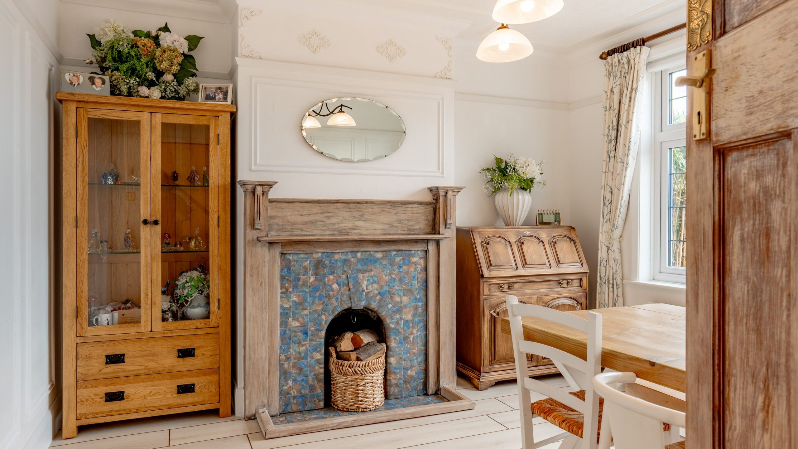 A cozy room with a wooden cabinet, ornate fireplace with blue tiles, and a wood floor. A round mirror and floral arrangement adorn the mantel. Nearby, a wooden dining table with chairs sits by a window with patterned curtains, and a vintage desk stands nearby.
