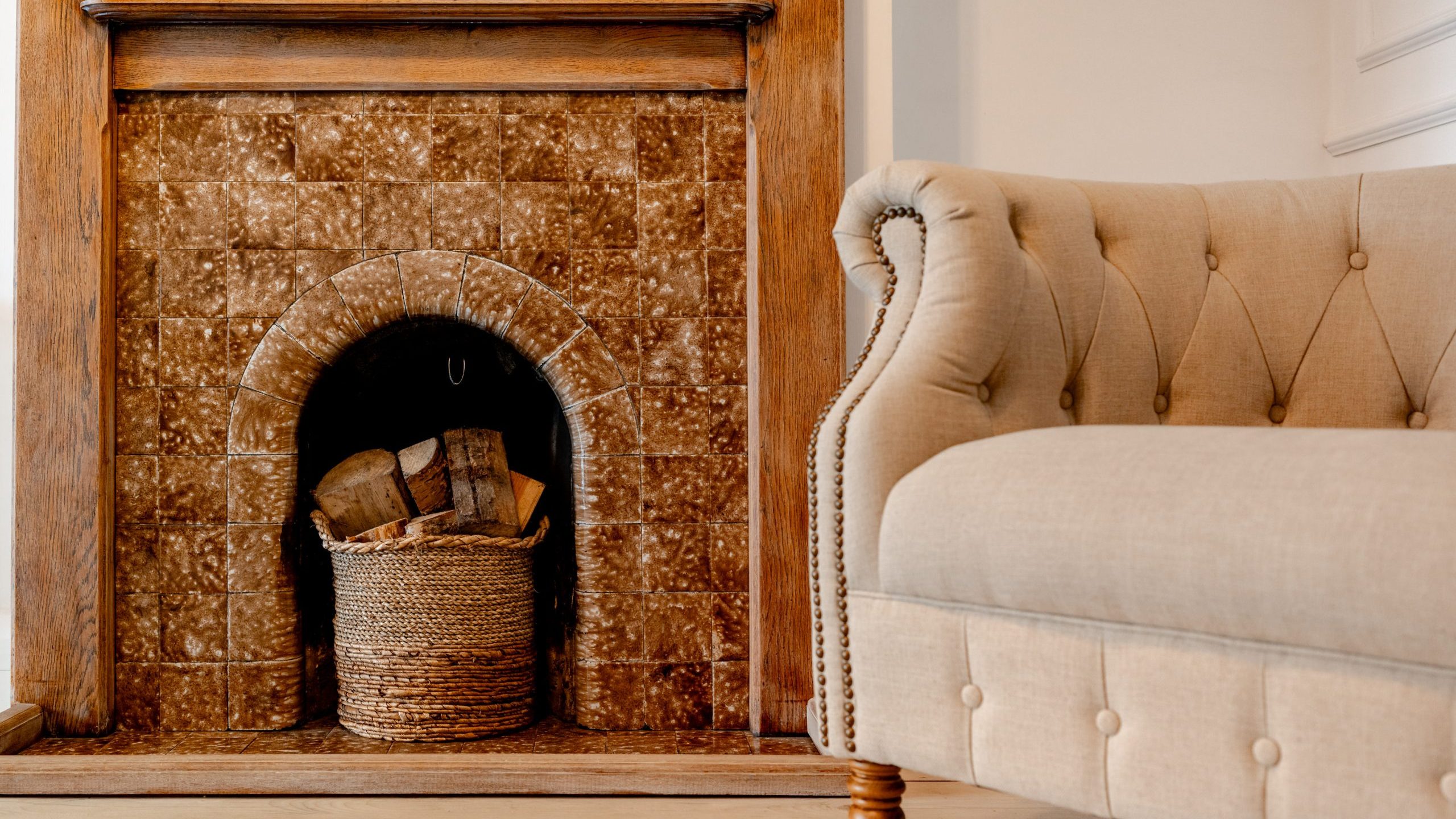 A cozy living room scene featuring a beige tufted sofa next to a wooden fireplace with brown tile. Inside the fireplace, there's a woven basket holding firewood, adding a rustic touch to the warm, inviting space.
