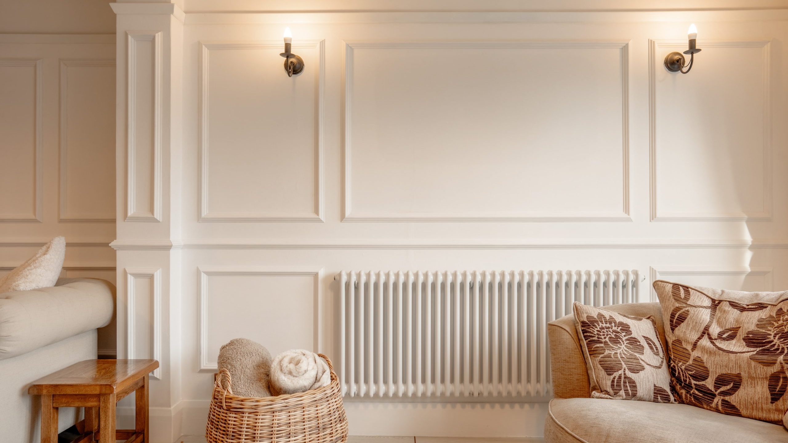 A cozy living room with a white paneled wall, two wall sconces, a central radiator, and a wicker basket with blankets. A beige sofa with floral cushions is on the right, and a small wooden stool is on the left.