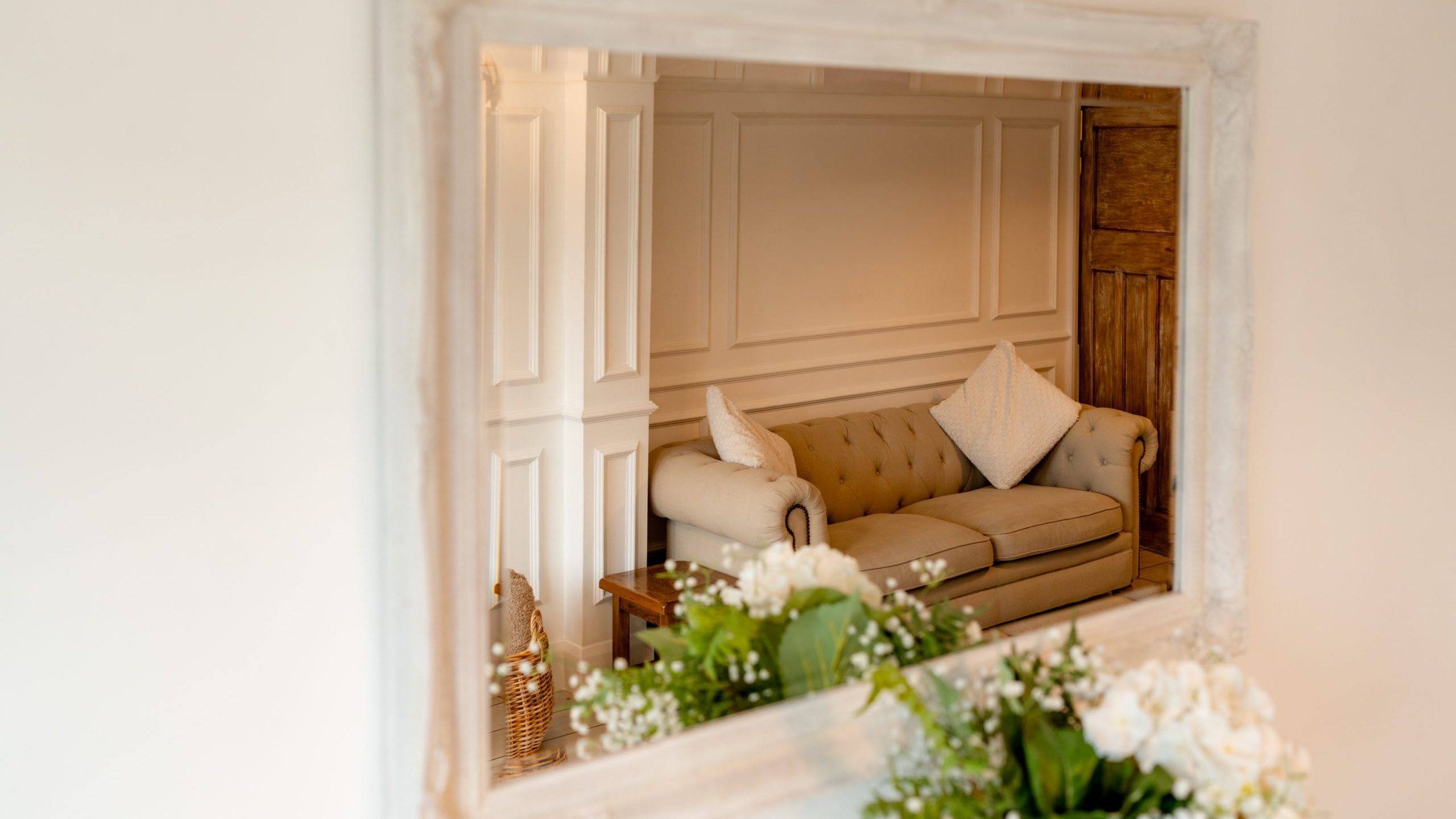 A large mirror reflects a cozy living room scene with a beige tufted sofa adorned with white pillows. The decor includes white paneled walls, a wooden door, and greenery with white flowers in the foreground.