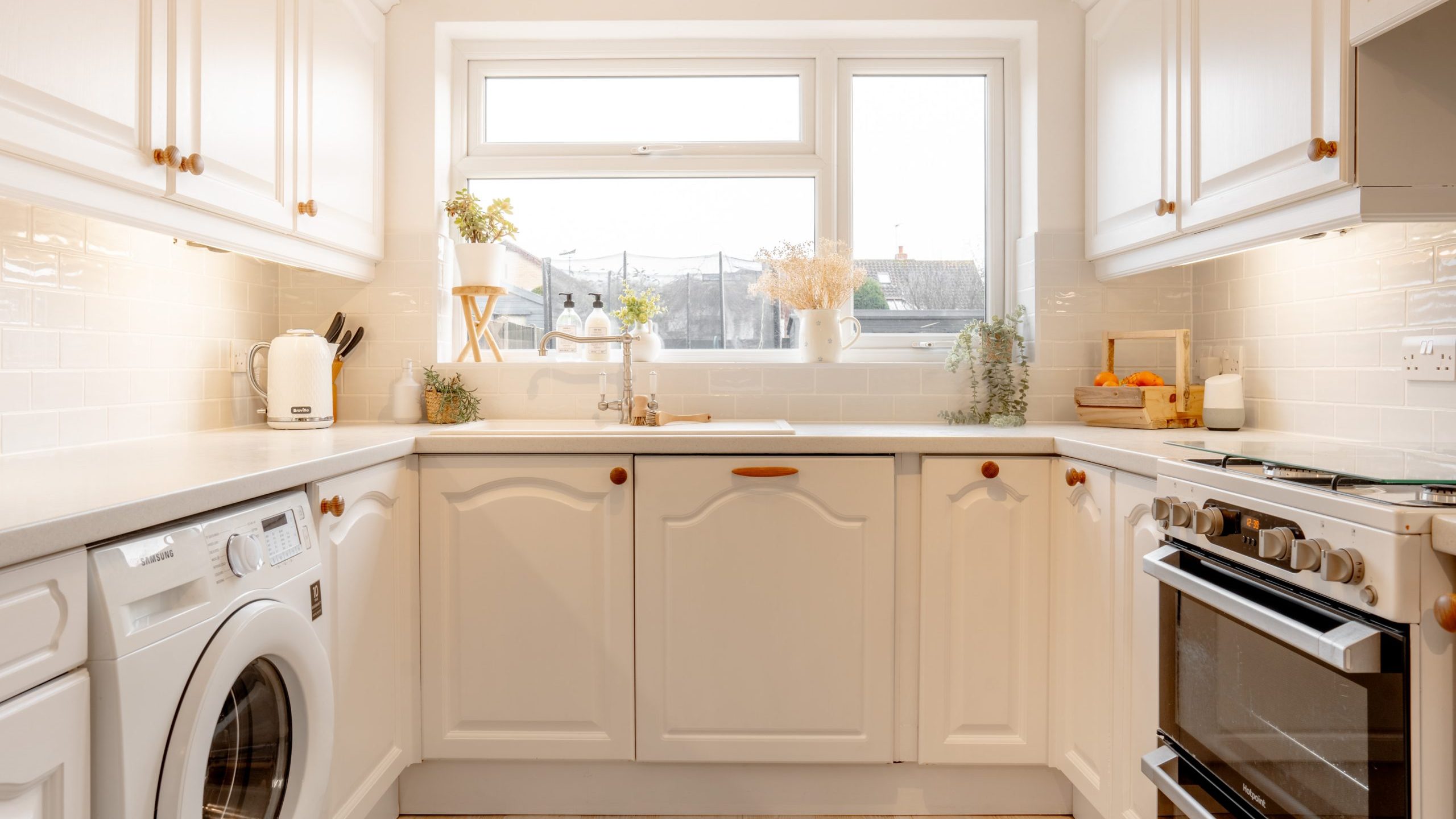A bright, modern kitchen featuring white cabinets, a washing machine, and a stovetop. Natural light streams through a large window with plants on the sill. The room has light wood flooring and subtle decorative elements.