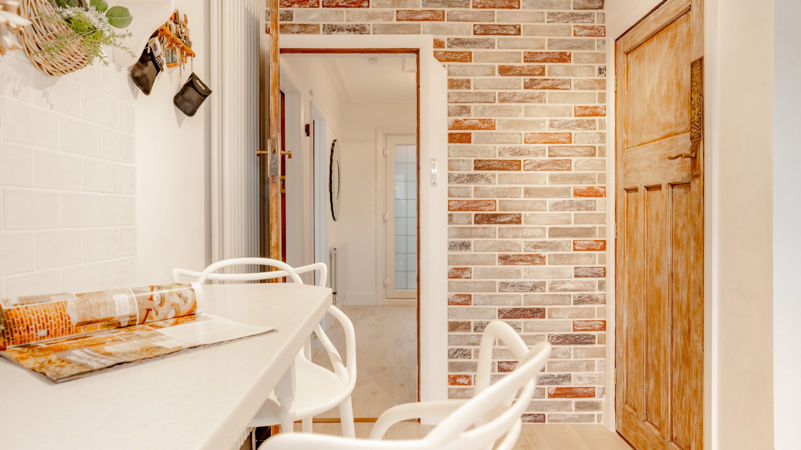 A cozy kitchen area with white bar stools at a countertop. The walls feature white tiles and an exposed brick design. A wooden door is on the right, and a doorway leads to a bright hallway with a mirror. Woven baskets hang on the wall.