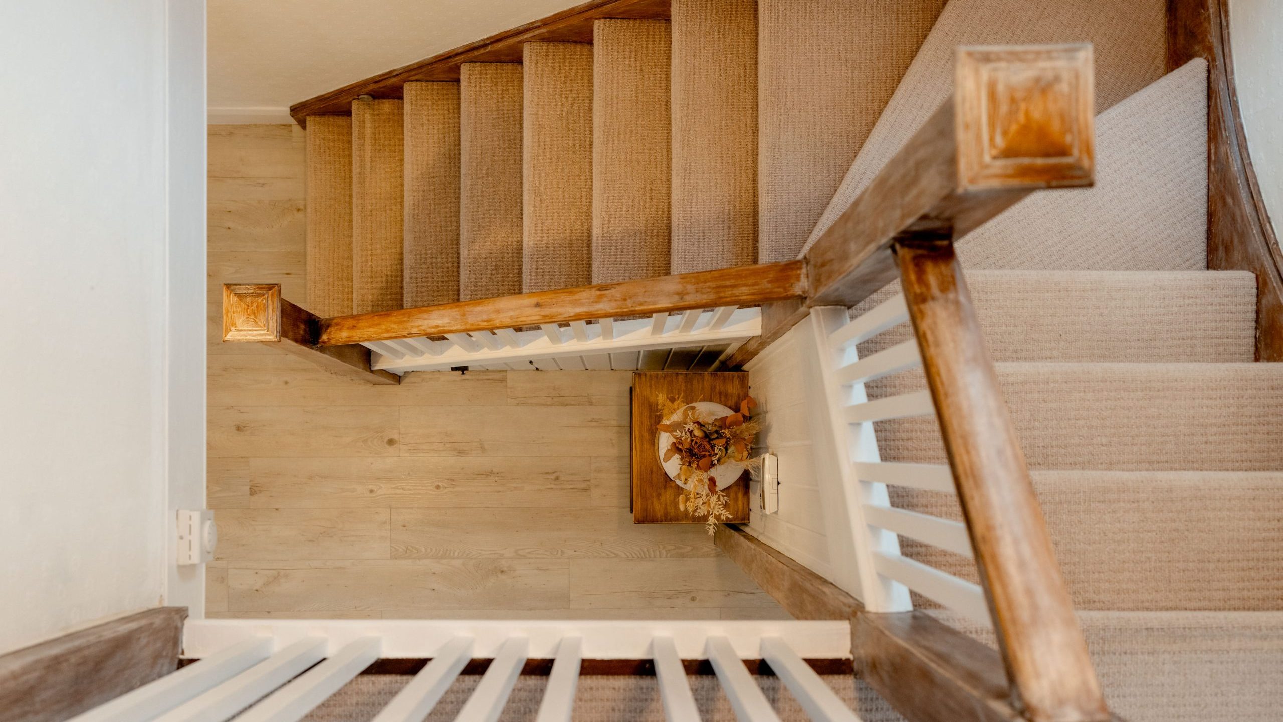 Top-down view of a wooden staircase with beige carpet, featuring a landing area with a decorative basket of dried flowers. The stairs have a wooden balustrade with white vertical slats, and the floor is light wood.