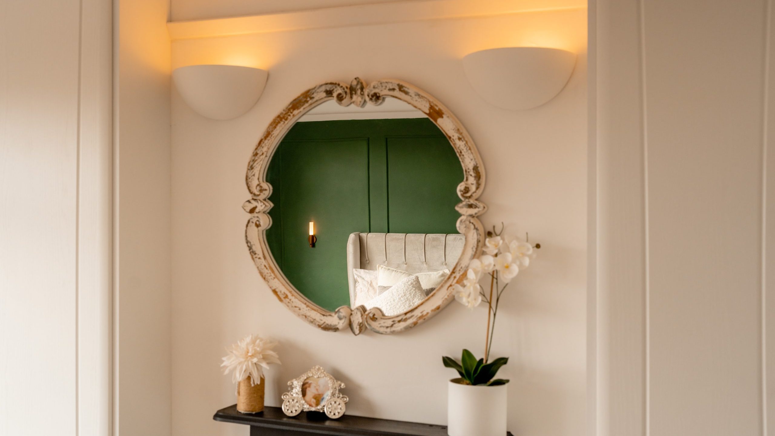 A vintage oval mirror hangs on a white wall, reflecting a green-painted room with a bed. Below the mirror, a black shelf holds a potted orchid and decorative items. Soft ambient lighting highlights the elegant, serene decor.