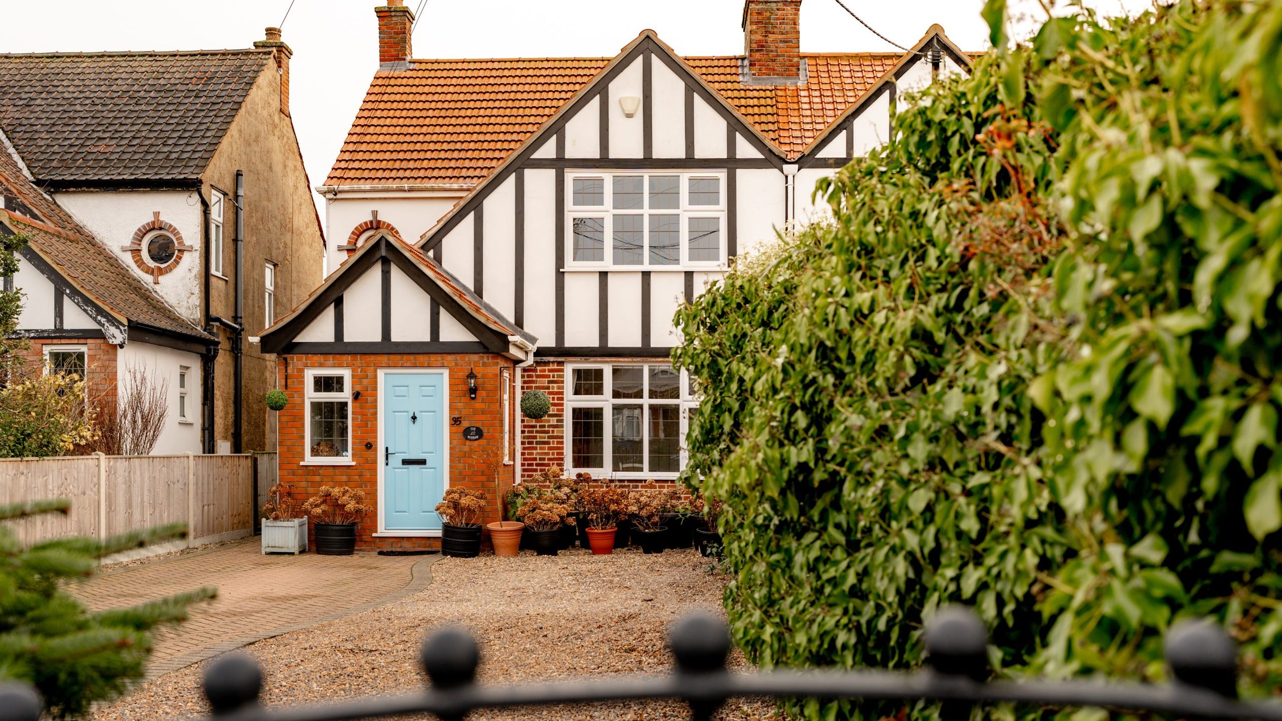 A charming two-story Tudor-style house with a gabled roof, white and black timber framing, and a bright blue front door. The front yard has potted plants, gravel, and hedges, with neighboring houses on either side.
